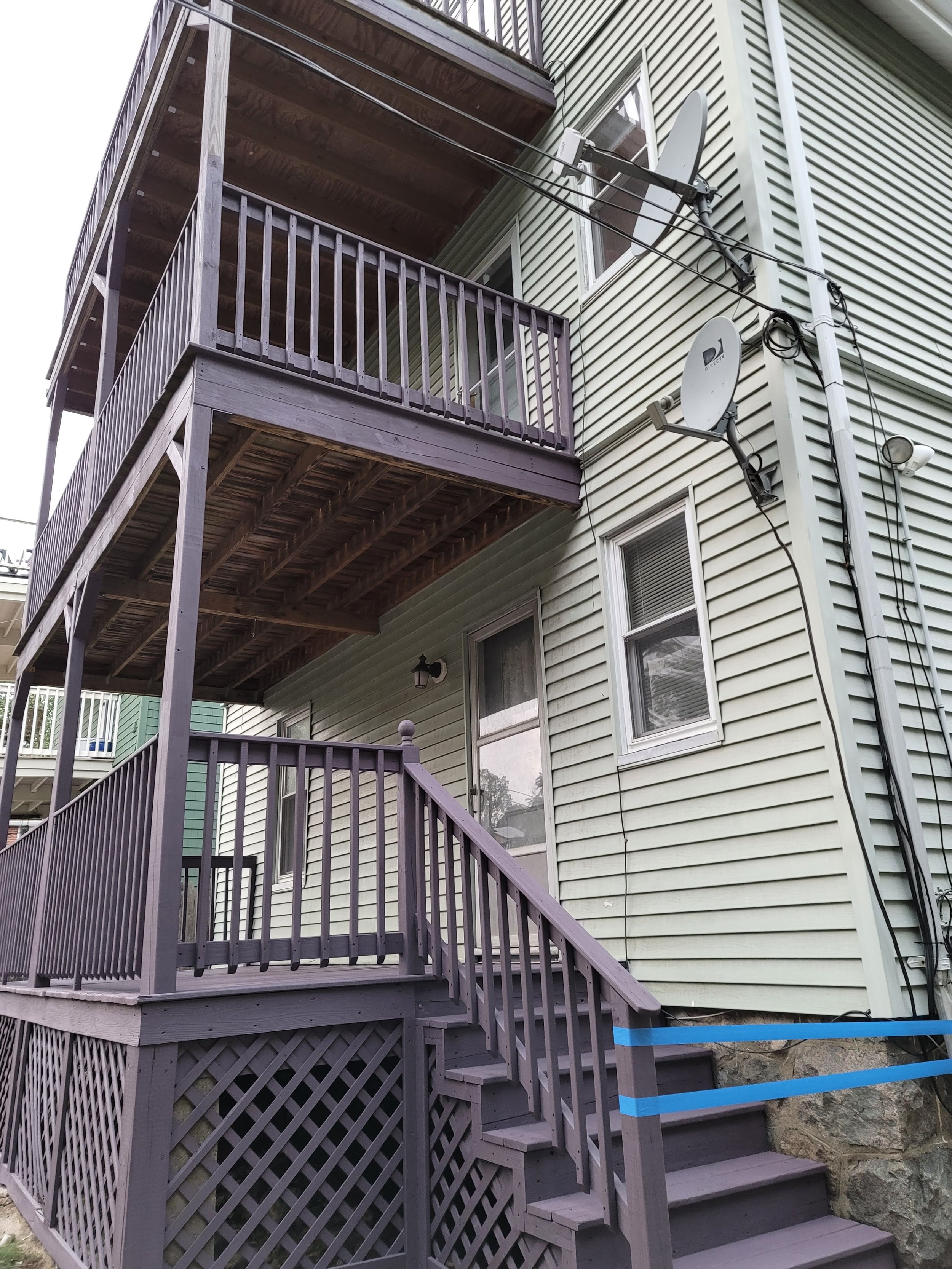 Exterior view of a two family house in Roslindale with two newly deck stained wooden balconies, satellite dishes, and pale green exterior siding.
