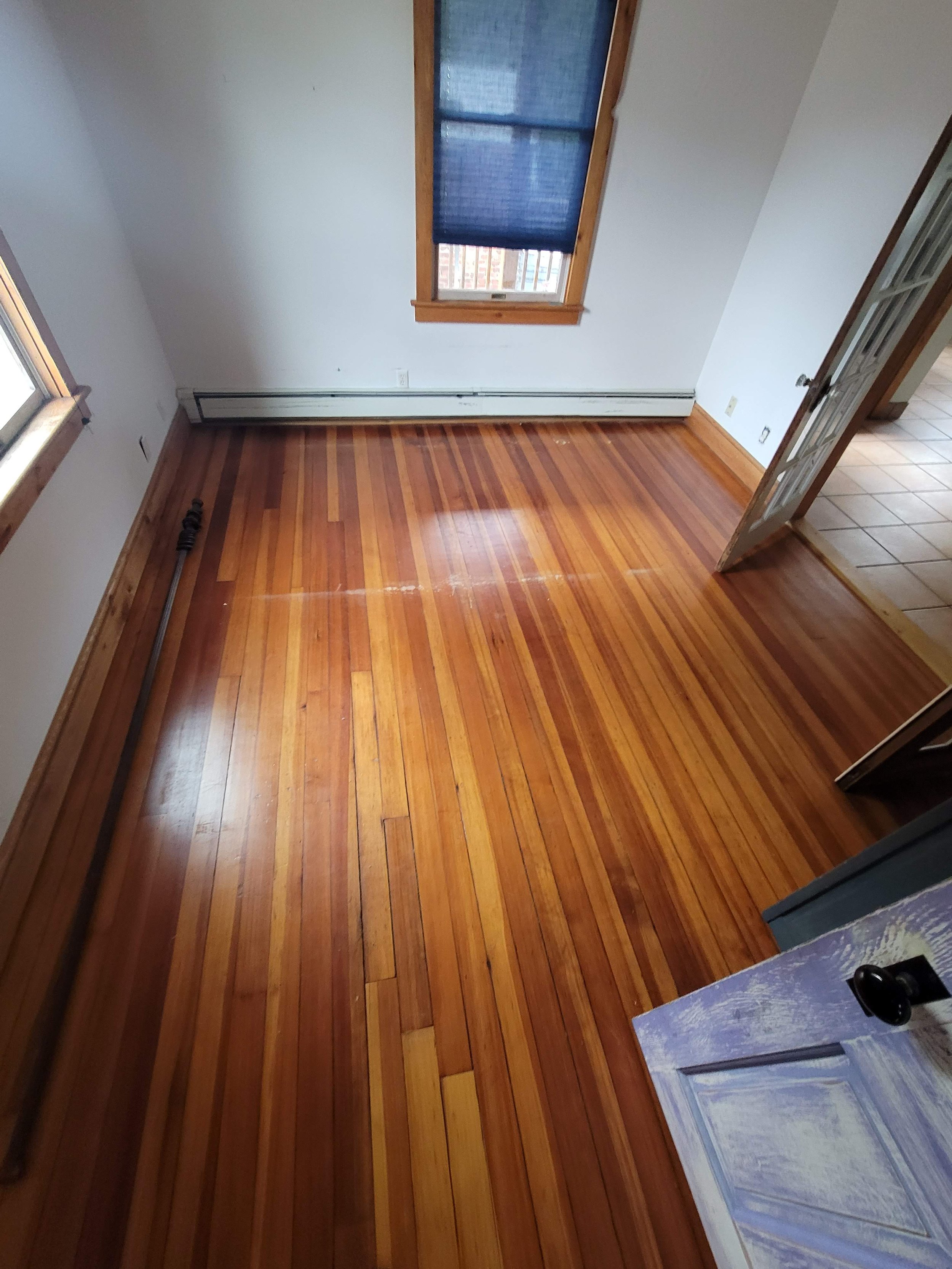 Empty room with wooden floors, white walls, two windows with blue blinds, and an open door leading to tiled flooring in Jamaica Plain, Boston.