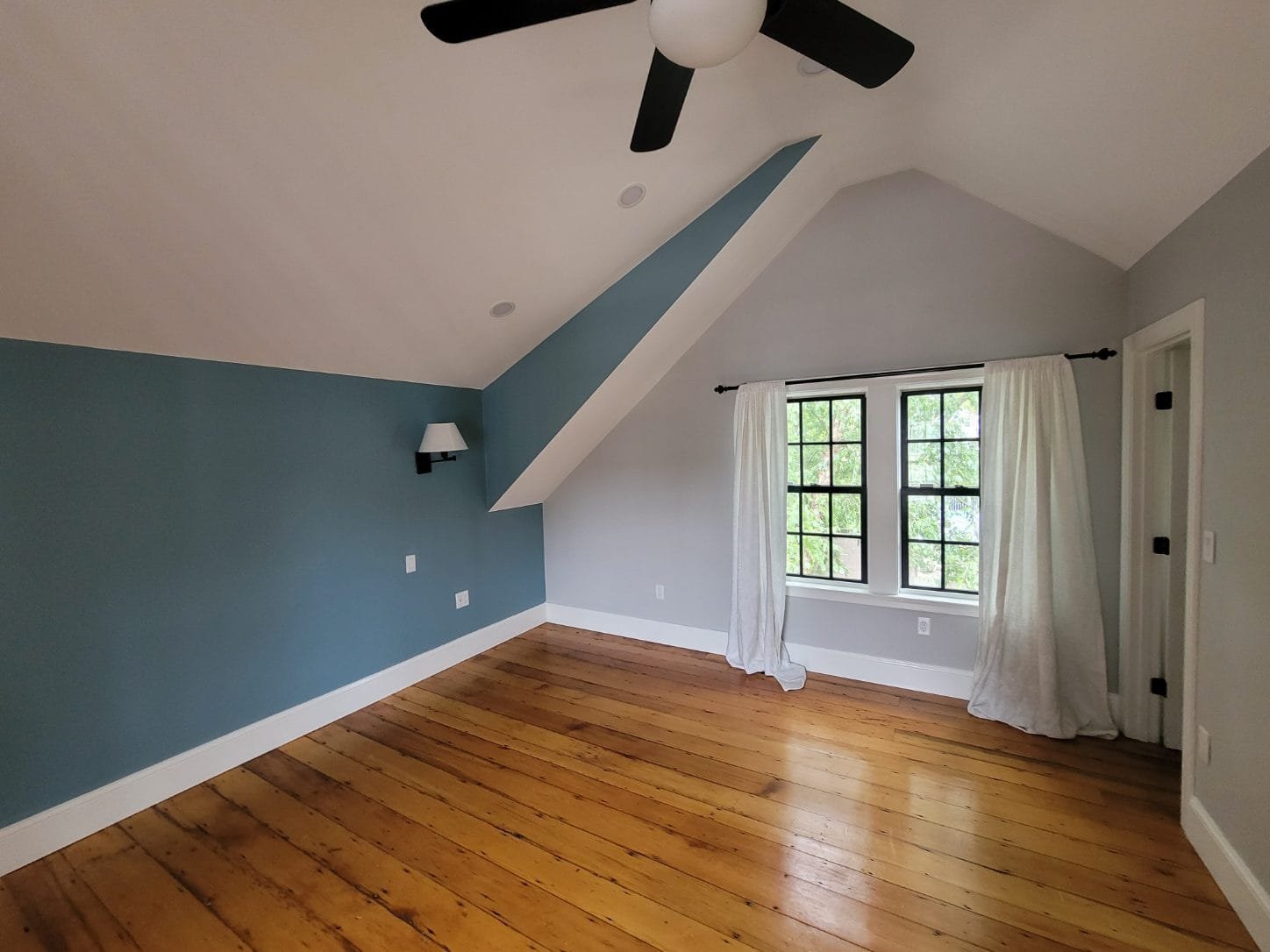 Empty room in a colonial style home in Dorchester, with wooden floor, blue and white walls, a window with white curtains, a ceiling fan, and a wall-mounted light fixture.