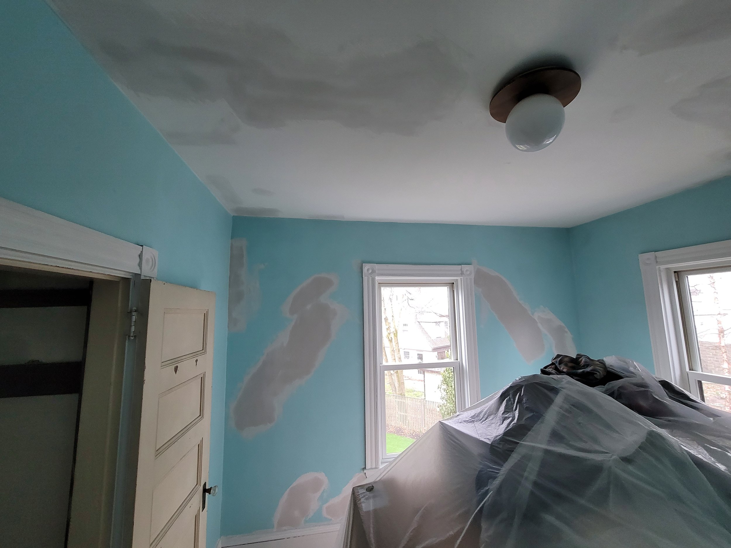 Room undergoing horse hair plaster repair on Patten Street, Roslindale with patches of joint compound on light blue walls and a covered piece of furniture.