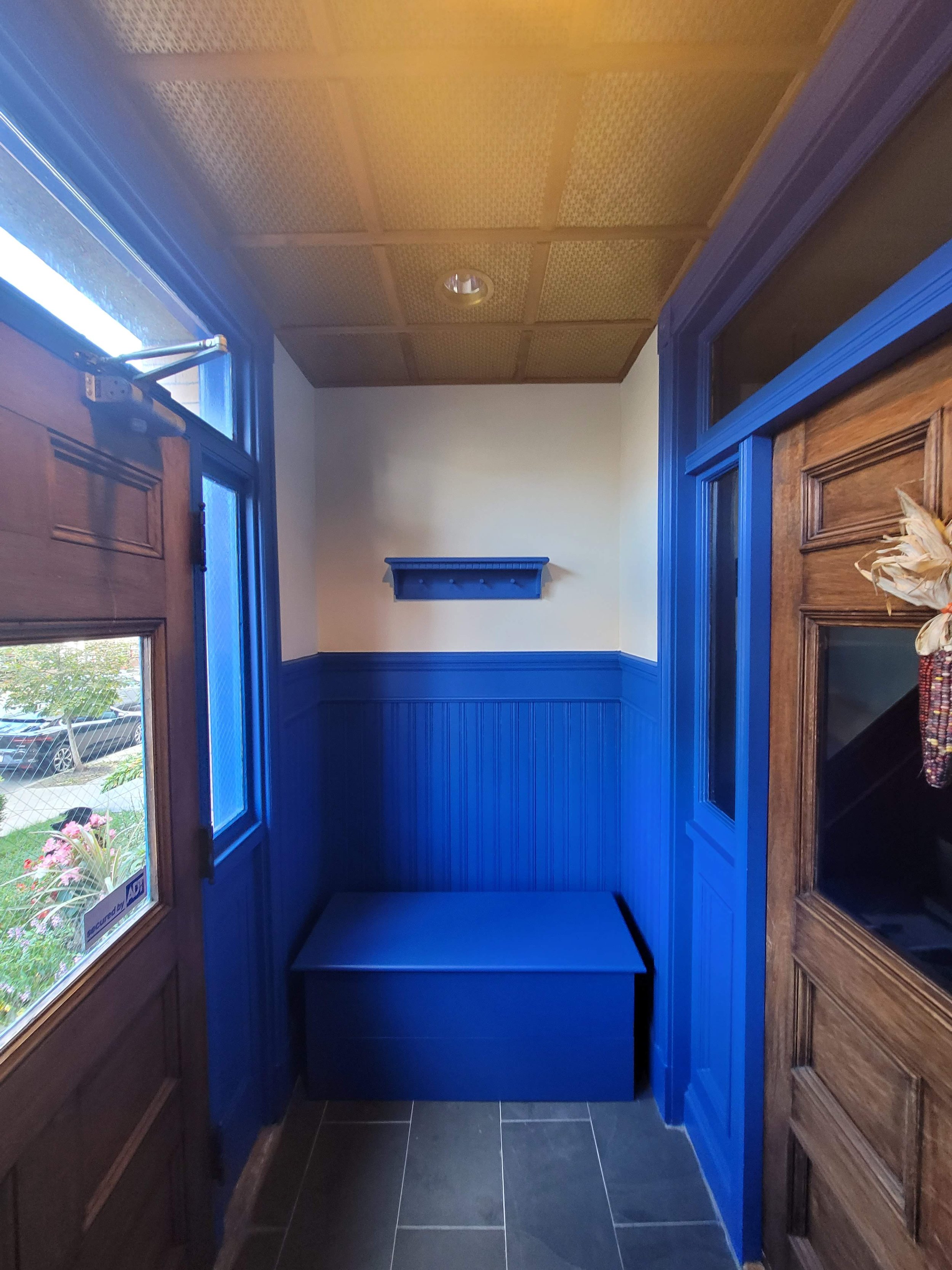 Corner of a foyer room on Jones Hill, Dorchester with wood and blue painted walls, wooden door, window, a small blue shelf, and a blue bench.