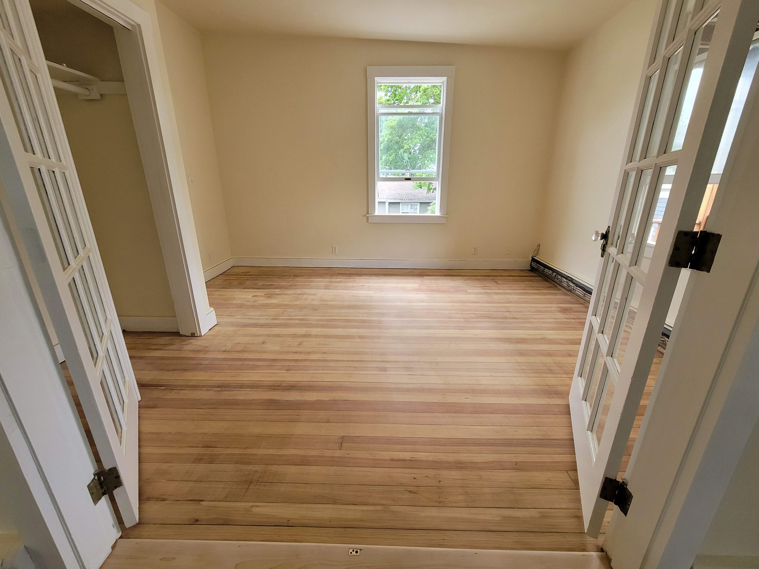 Empty room with recently refinished by Ryan Painting hardwood floor, white walls, and a window showing green trees outside. French doors open into the room.
