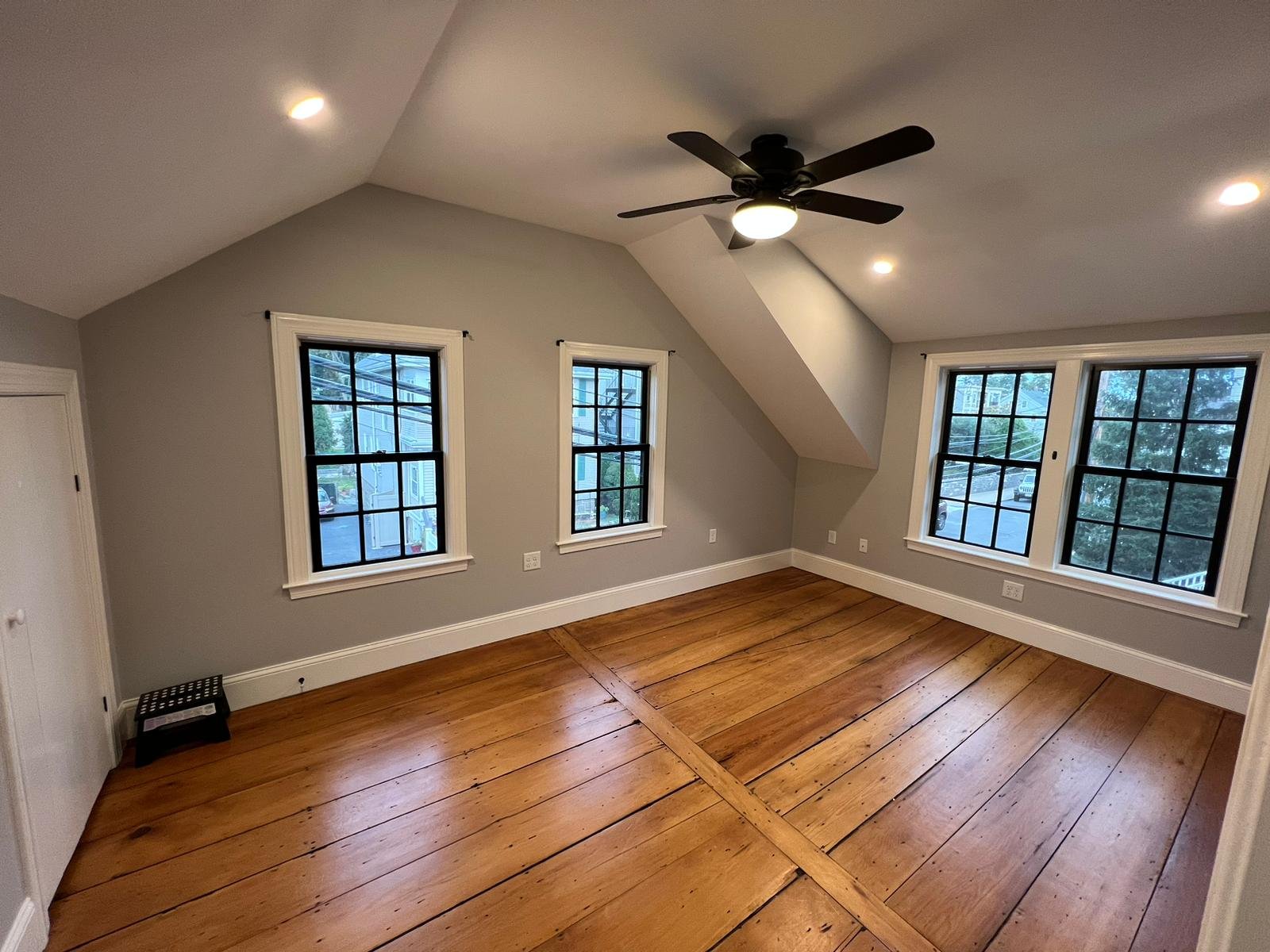 Empty room with hardwood floor, gray walls, four windows with black frames, ceiling fan, and recessed lighting recently painted by Ryan Painting in Dorchester, Boston.