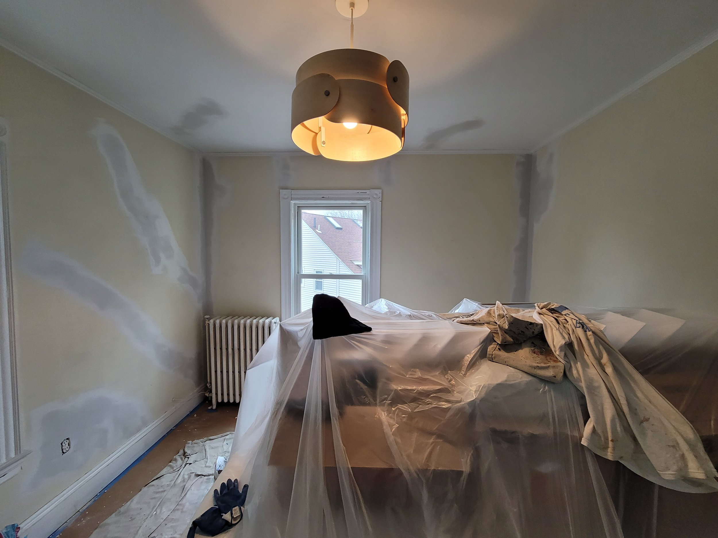 Room in a Victorian-style home in Boston with plaster and lathe walls being patched and painted, covered with plastic sheeting, with a rooftop visible through the window.