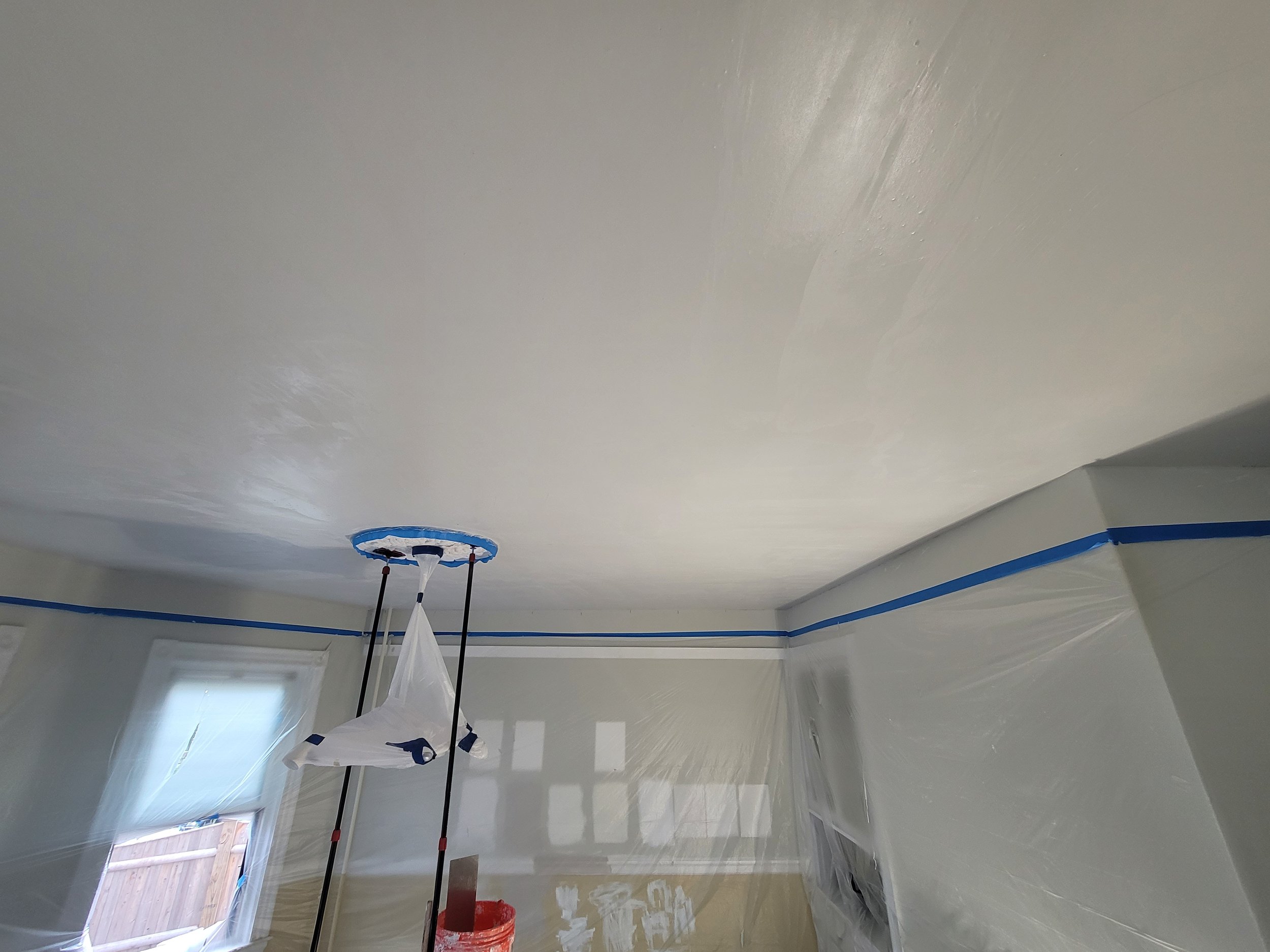 A room with recent ceiling repairs being painted white by Ryan Painting, covered in plastic sheeting, with a Ryan Painting crew member's pole and bucket visible, and ceiling light fixture covered with plastic.