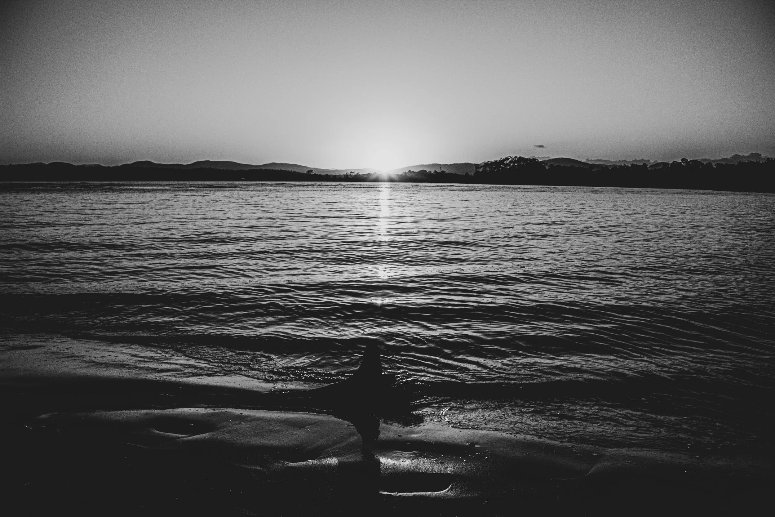 Black and white photo of a river with the sun setting behind distant hills. A person is sitting on the shoreline in the foreground, facing the water.