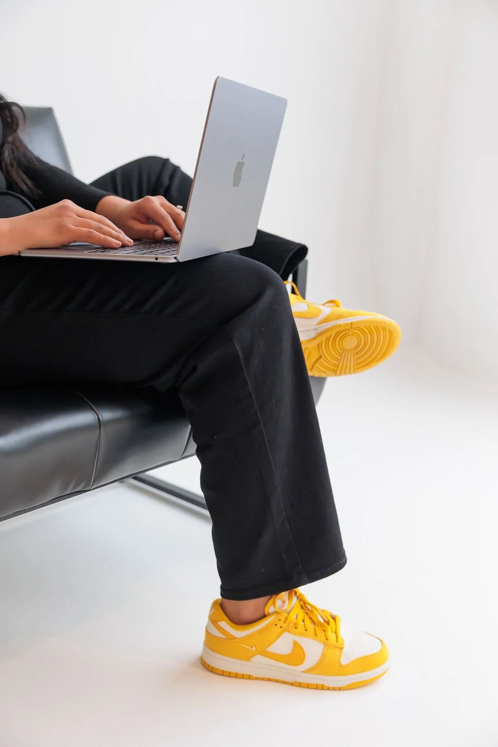 Bookkeeping and business coach for service based business owners. Person sitting on a black bench, using a silver MacBook laptop, wearing black pants and bright yellow Nike sneakers, with another yellow sneaker visible on the bench.