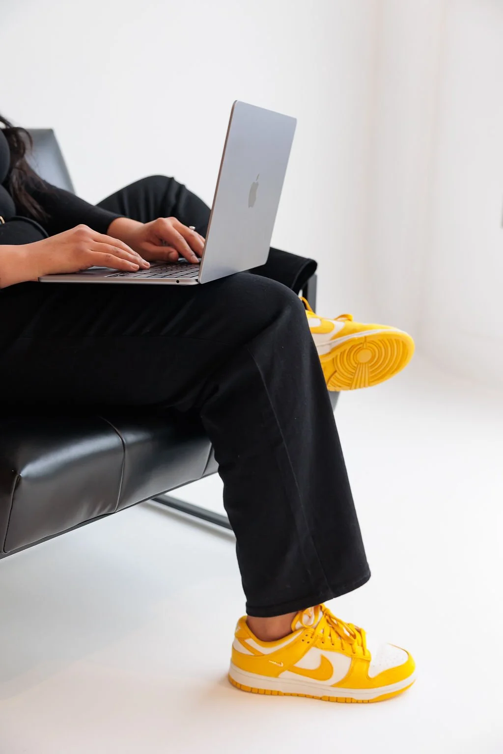 Person sitting on a black bench typing on a silver MacBook, wearing yellow and white sneakers, black pants, and a black top, in a white room.