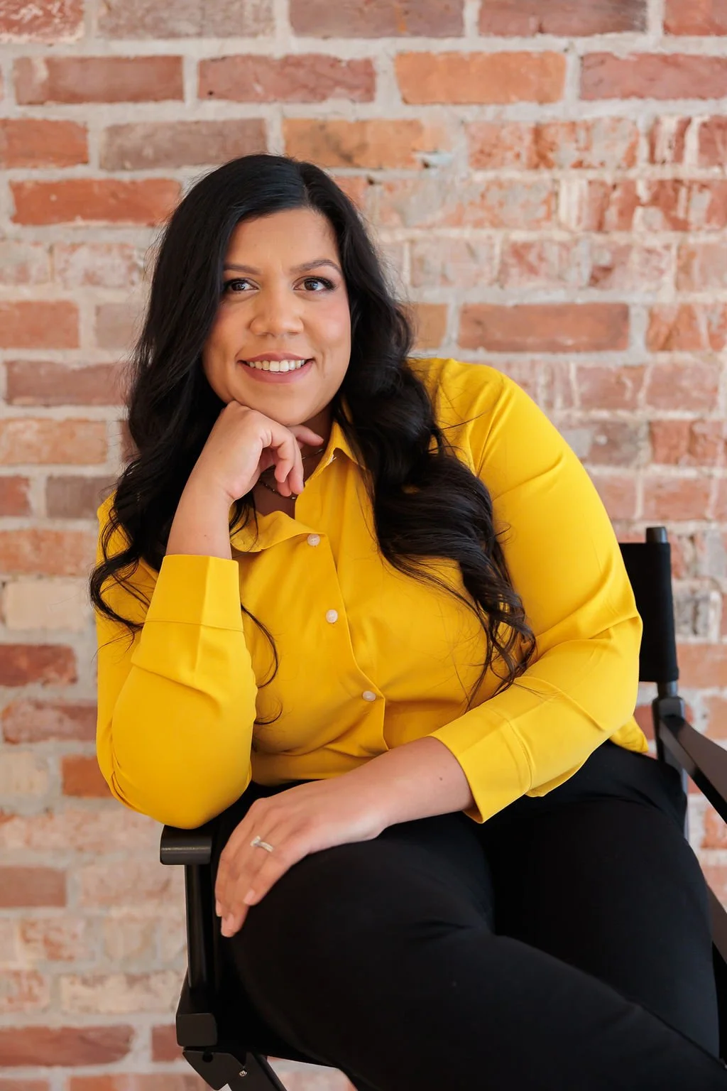 Bookkeeping and business coach for service based business owners. A woman with long black hair wearing a yellow shirt, sitting on a black chair, smiling, with a brick wall background.