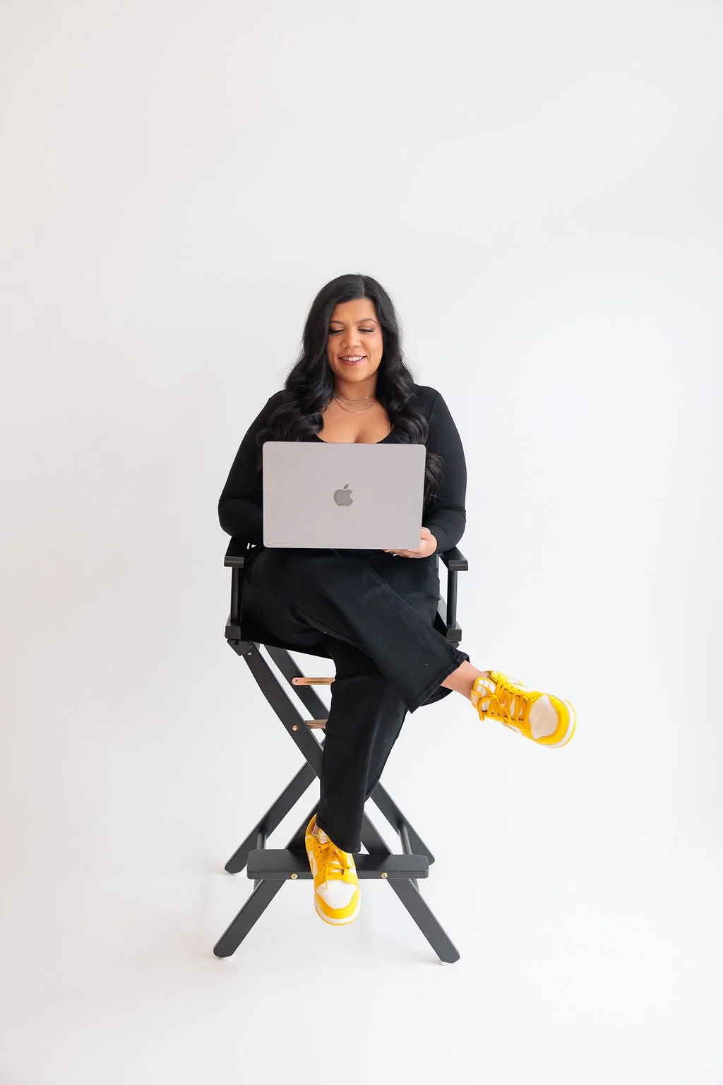 A woman with long dark hair, wearing a black top and black pants with yellow sneakers, sitting on a director's chair and using a silver MacBook against a plain white background.
