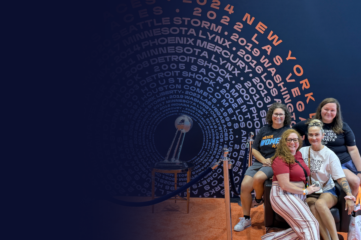 Four women sitting and standing in front of a large, spiral graphic mural with text and years, and a trophy displayed on a table beside them at an indoor event.