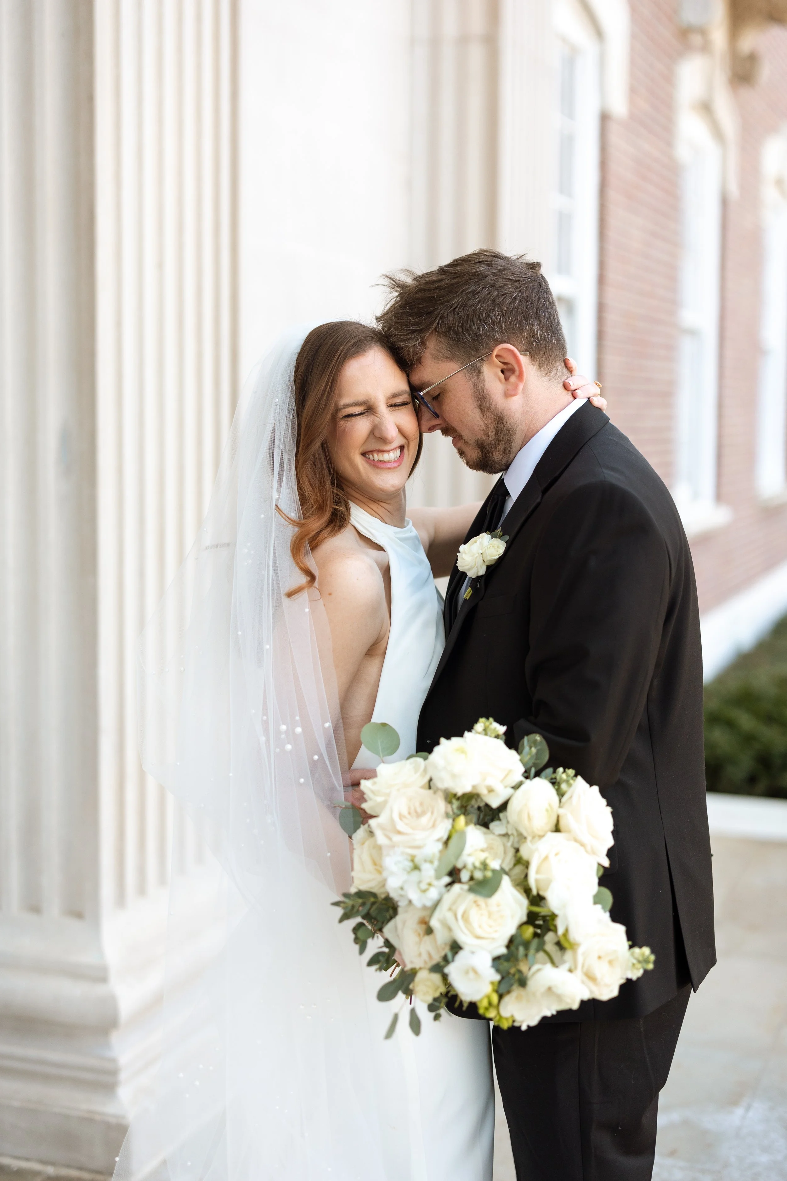 A bride and groom sharing an intimate moment outdoors, with the bride holding a large bouquet of white roses and greenery, against a background of a brick building and white column.