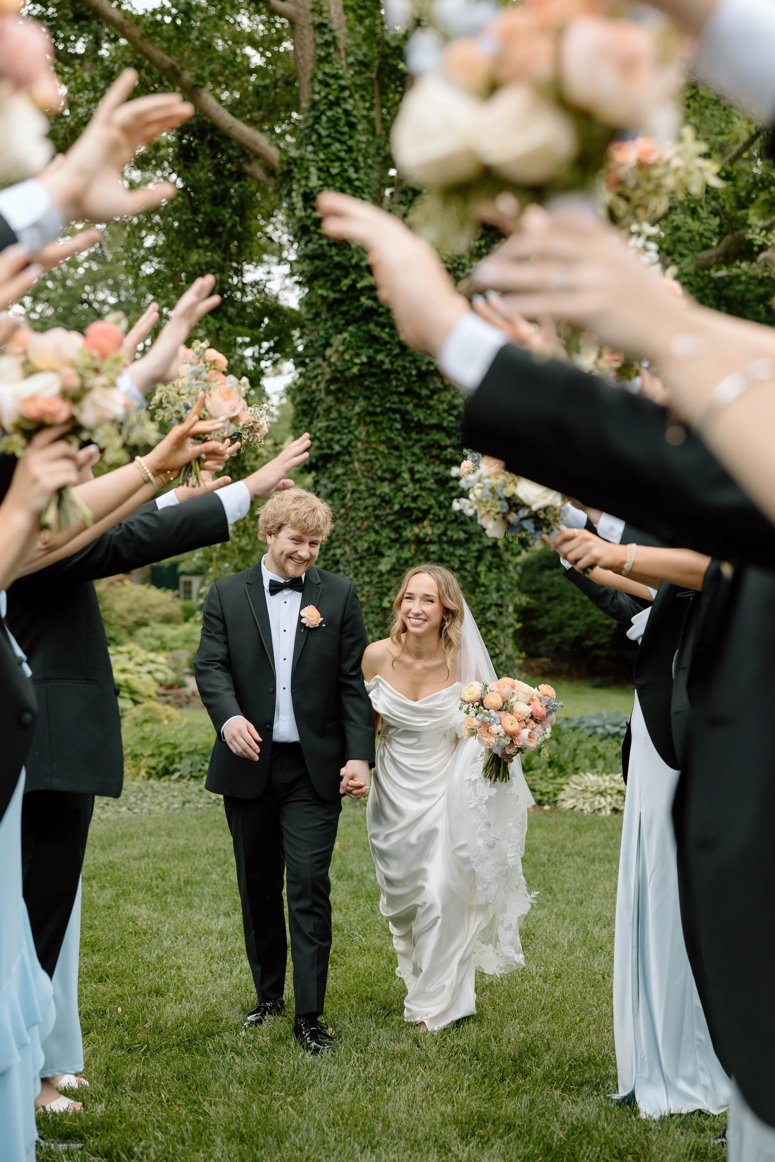 A newlywed couple walking hand-in-hand through a dashingly dressed wedding procession outdoors, with the bride holding a bouquet of peach and cream flowers, surrounded by friends and family in formal attire, celebrating their wedding day.