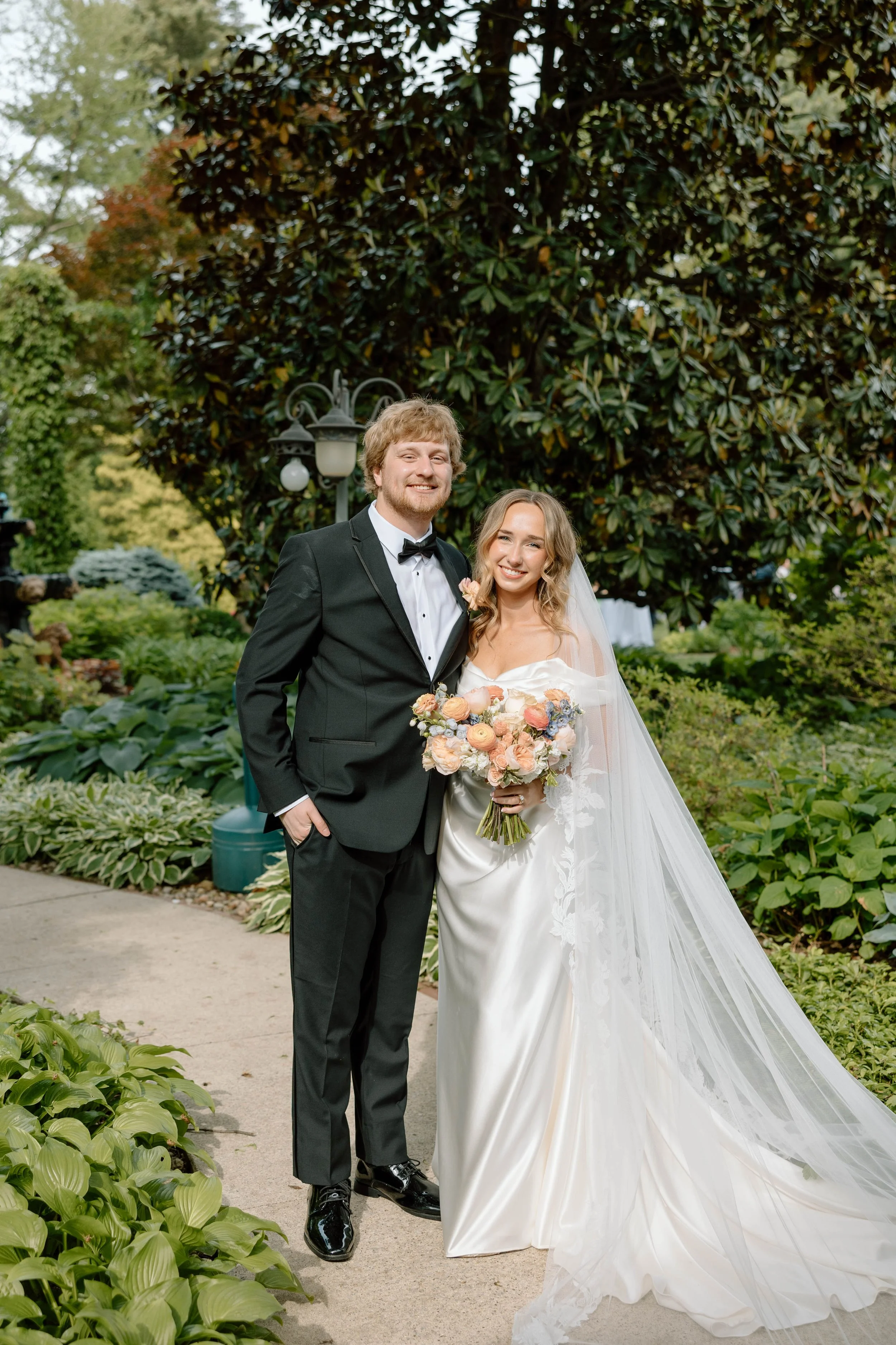 A newlywed couple standing outdoors in a garden, smiling at the camera, with lush greenery and trees in the background. The bride wears a white wedding gown with a long veil and holds a colorful bouquet, while the groom wears a black tuxedo with a bow tie.