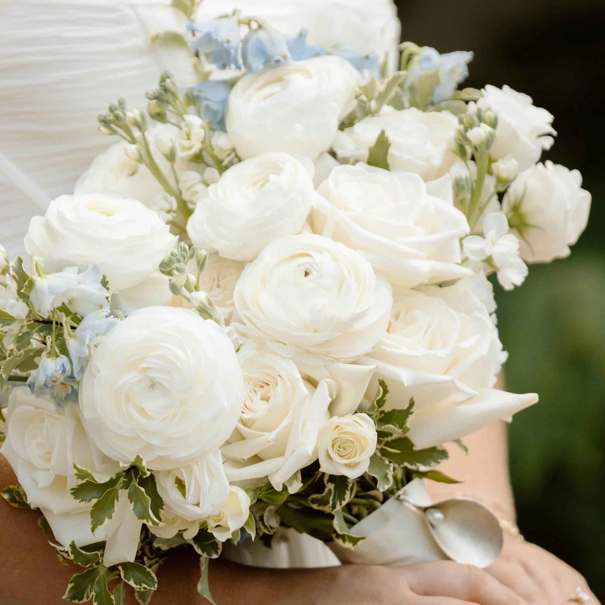 A wedding bouquet featuring white roses, ranunculus, calla lilies, and small blue flowers held by a bride.