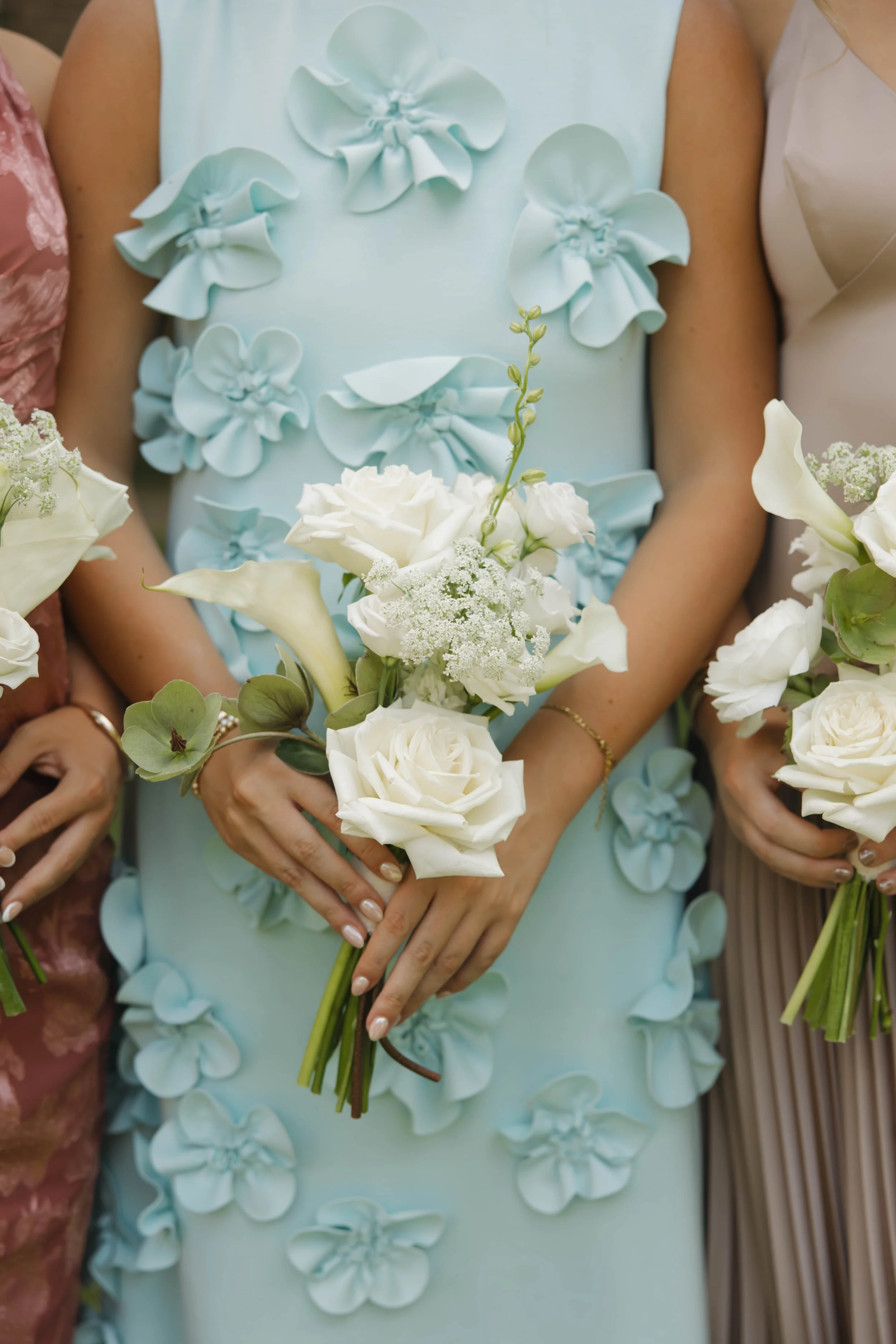 Close-up of women holding white floral bouquets at a wedding or celebration, with women wearing pastel dresses with floral embellishments.