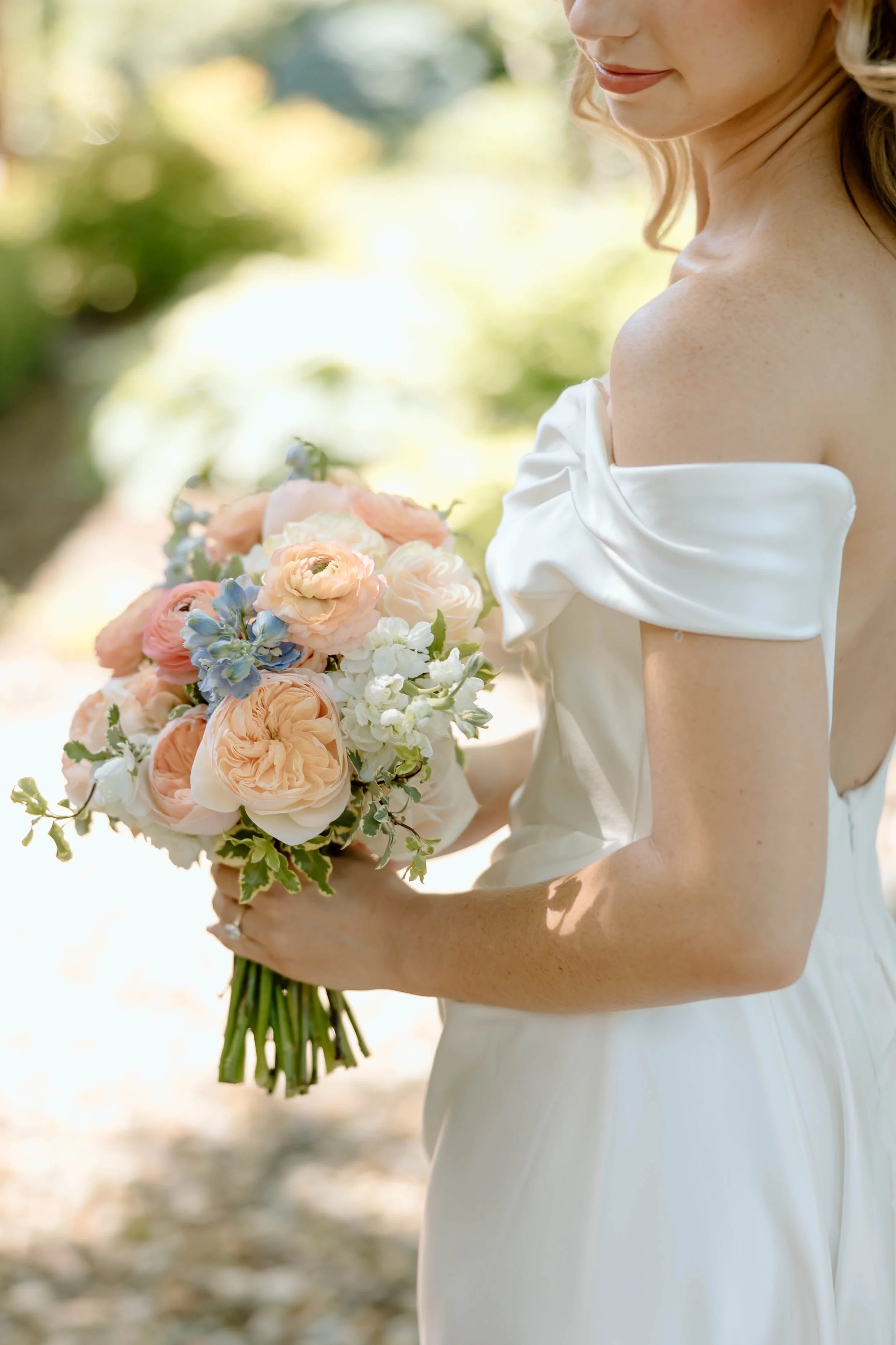 A bride in a white off-shoulder wedding dress holding a bouquet of pink, white, and blue flowers outdoors.