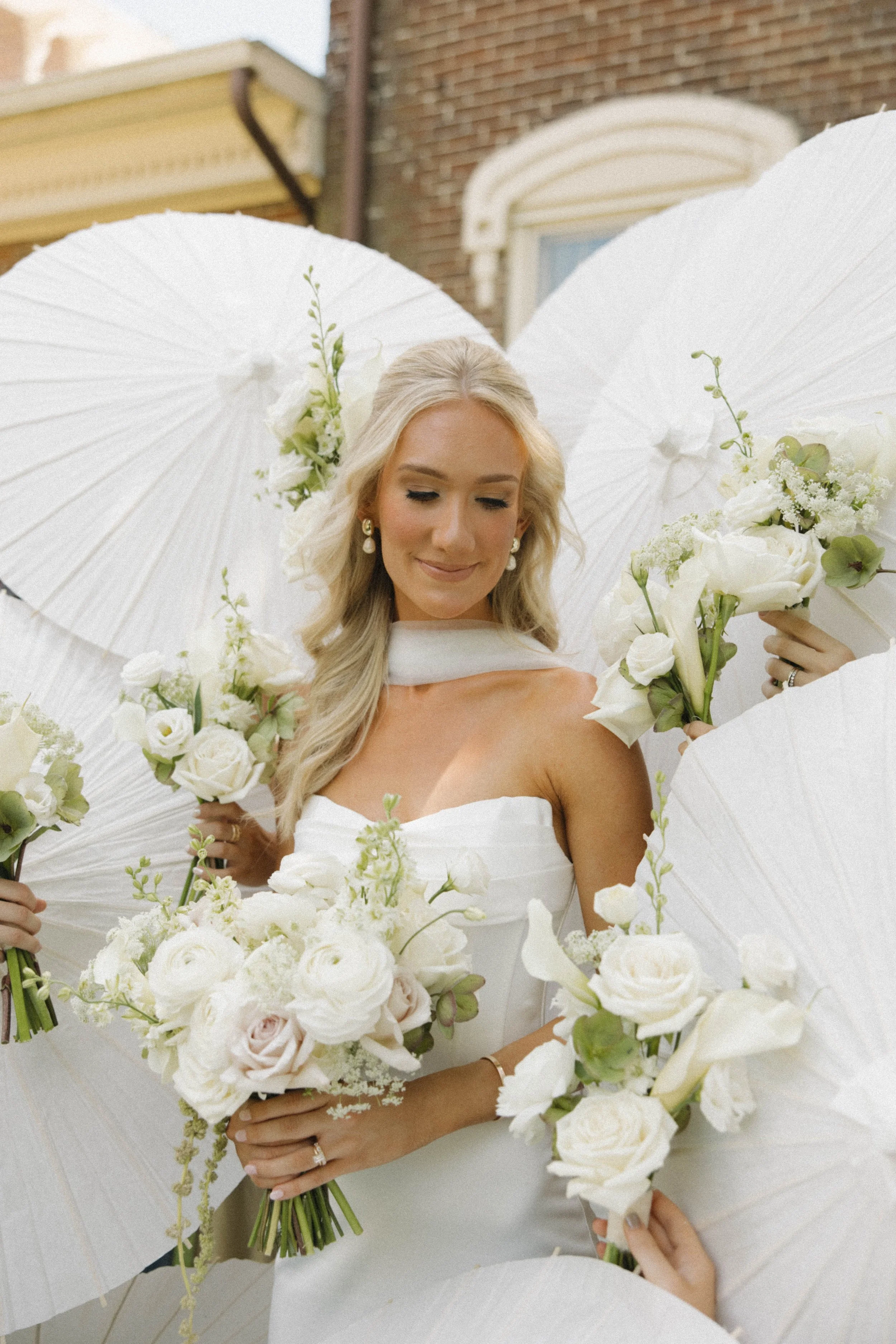 Bride in white dress holding a bouquet of white flowers, surrounded by white umbrellas, smiling with her eyes closed outdoors.