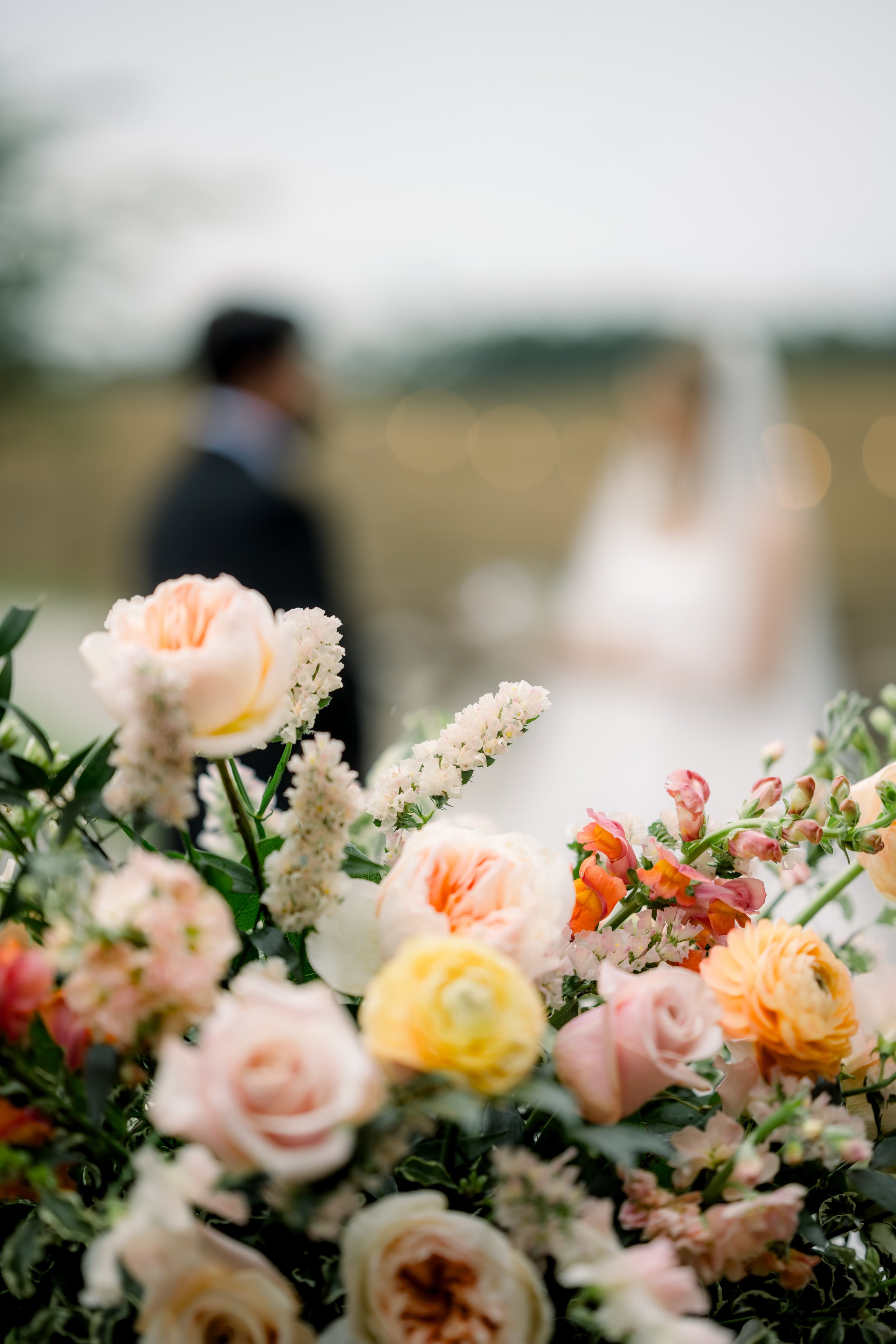 Blurred image of a wedding scene with a bride and groom in the background and a bouquet of pastel-colored flowers in the foreground.