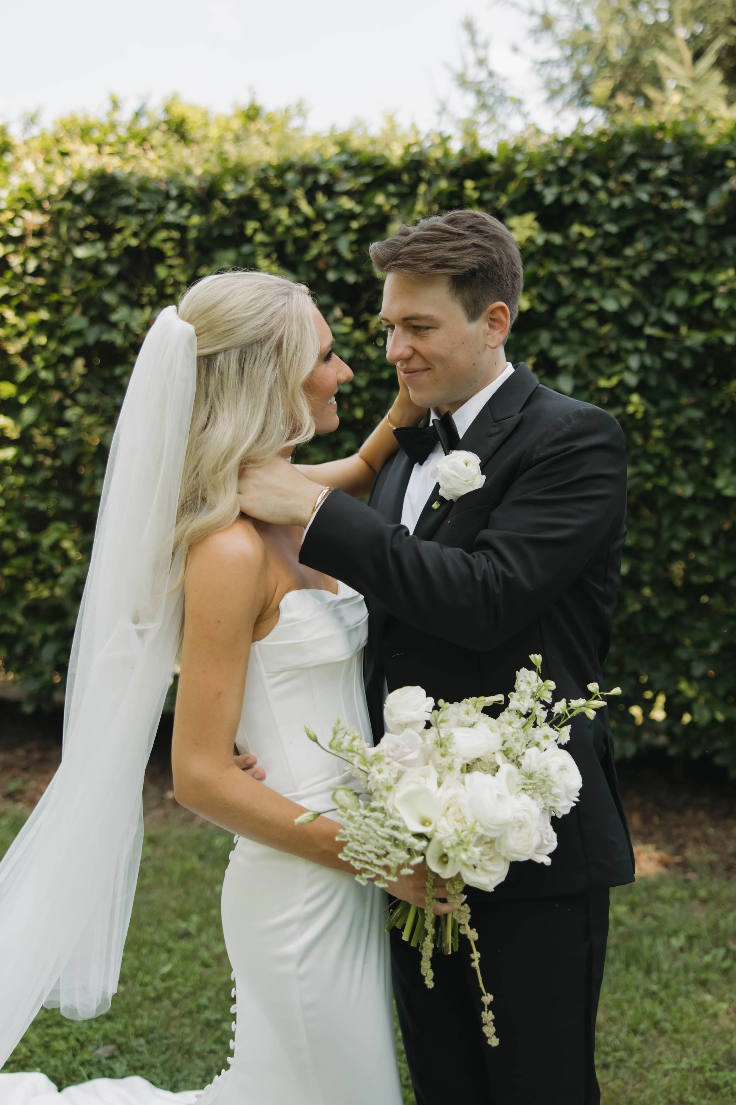 A bride and groom share a moment outdoors, with the bride holding a bouquet of white flowers, as the groom gently holds her neck.