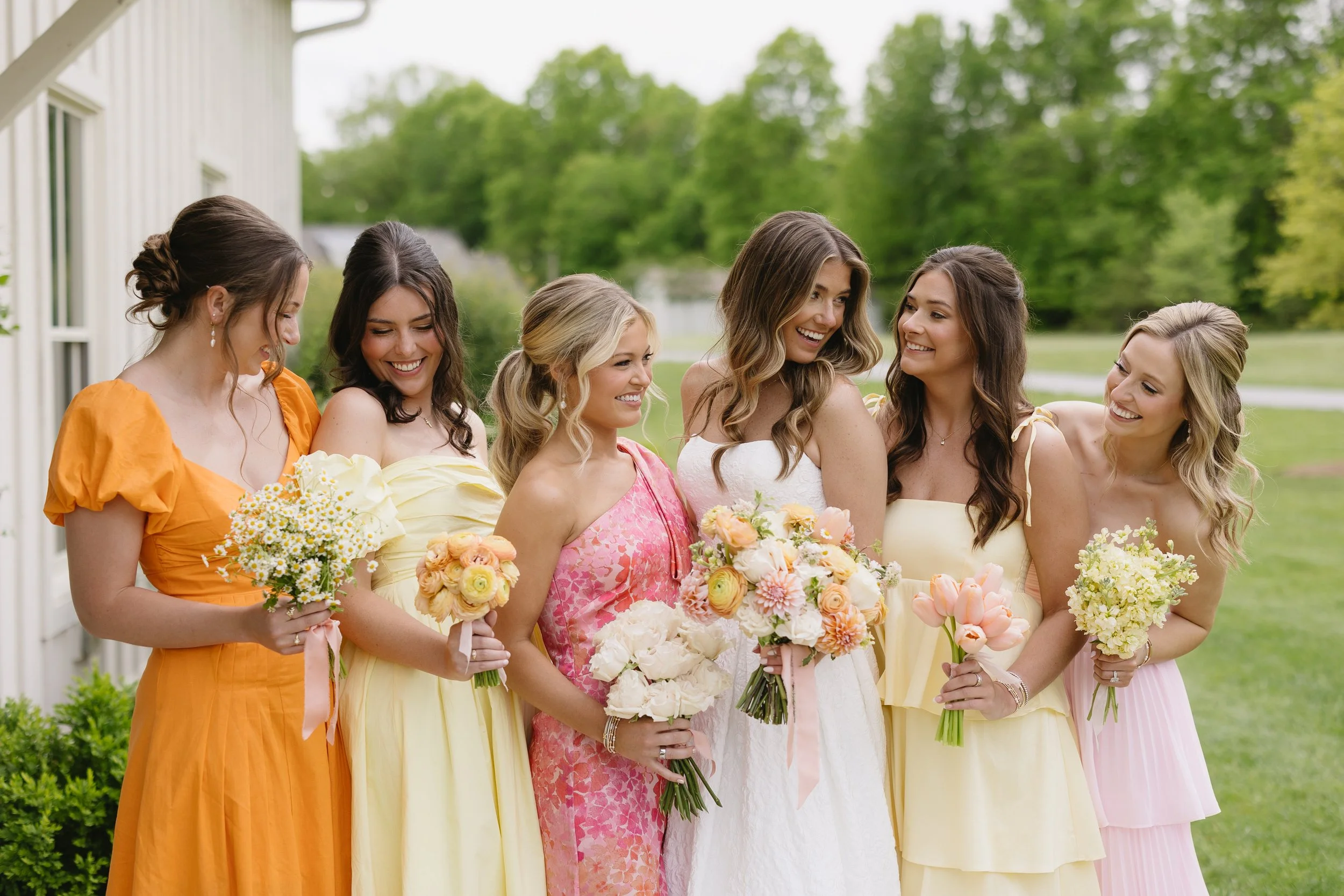 A group of seven women, including the bride in a white dress, standing outdoors on a grassy field with trees in the background. They are dressed in pastel and colorful dresses and holding bouquets of flowers, smiling and enjoying each other's company
