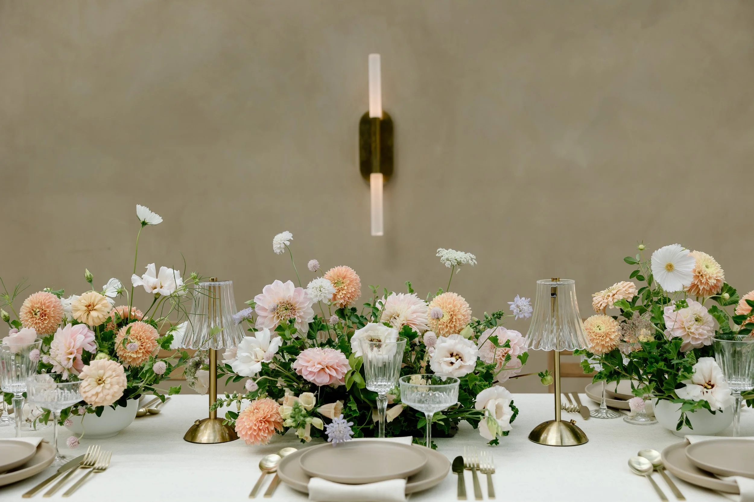 Elegant table setting with a floral centerpiece, candles, and tableware on a white tablecloth, with a neutral beige wall and modern wall sconce in the background.