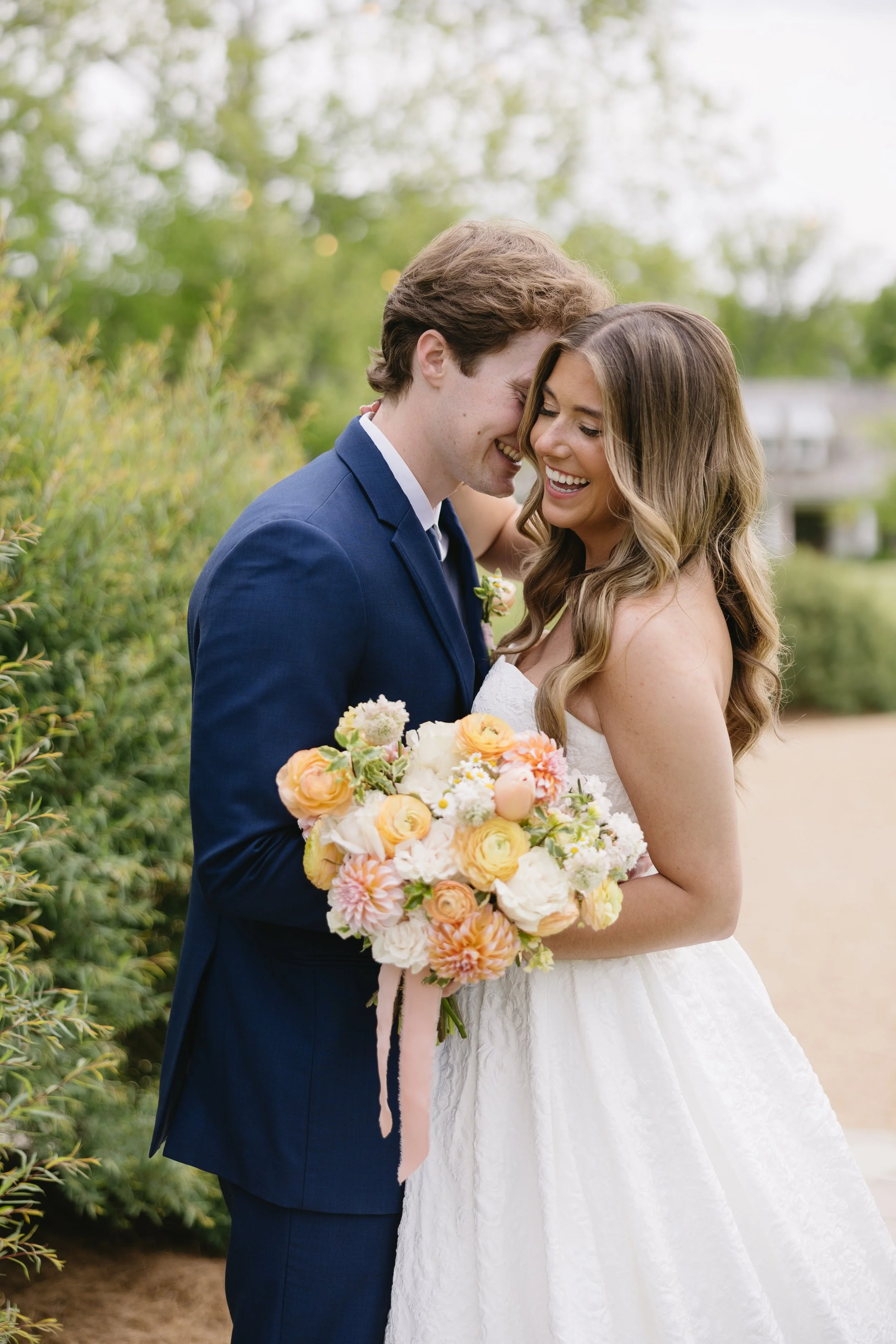 A newlywed couple sharing a joyful moment outdoors; the bride is holding a bouquet of colorful flowers, and they are smiling with their foreheads touching.