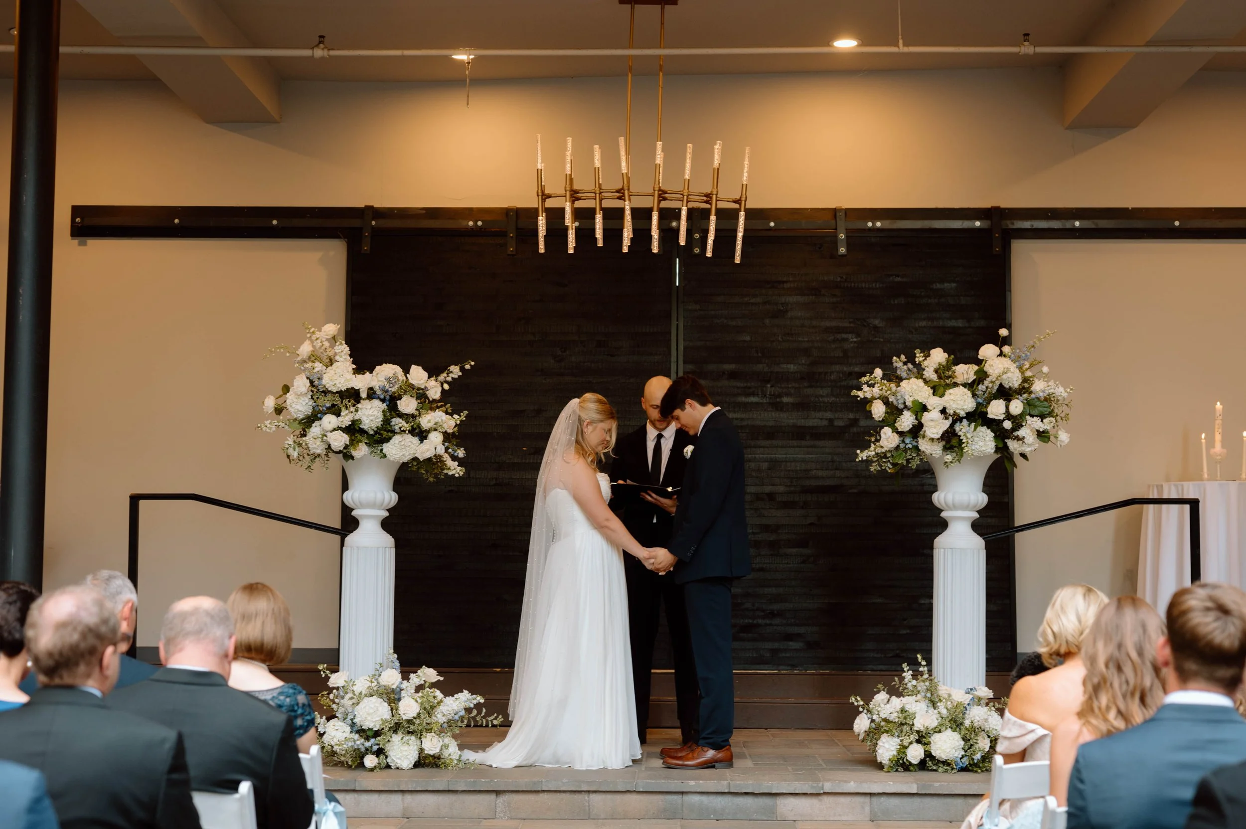 A bride and groom holding hands and facing each other during their wedding ceremony, with an officiant standing behind them, in a decorated indoor venue with floral arrangements and a dark backdrop.