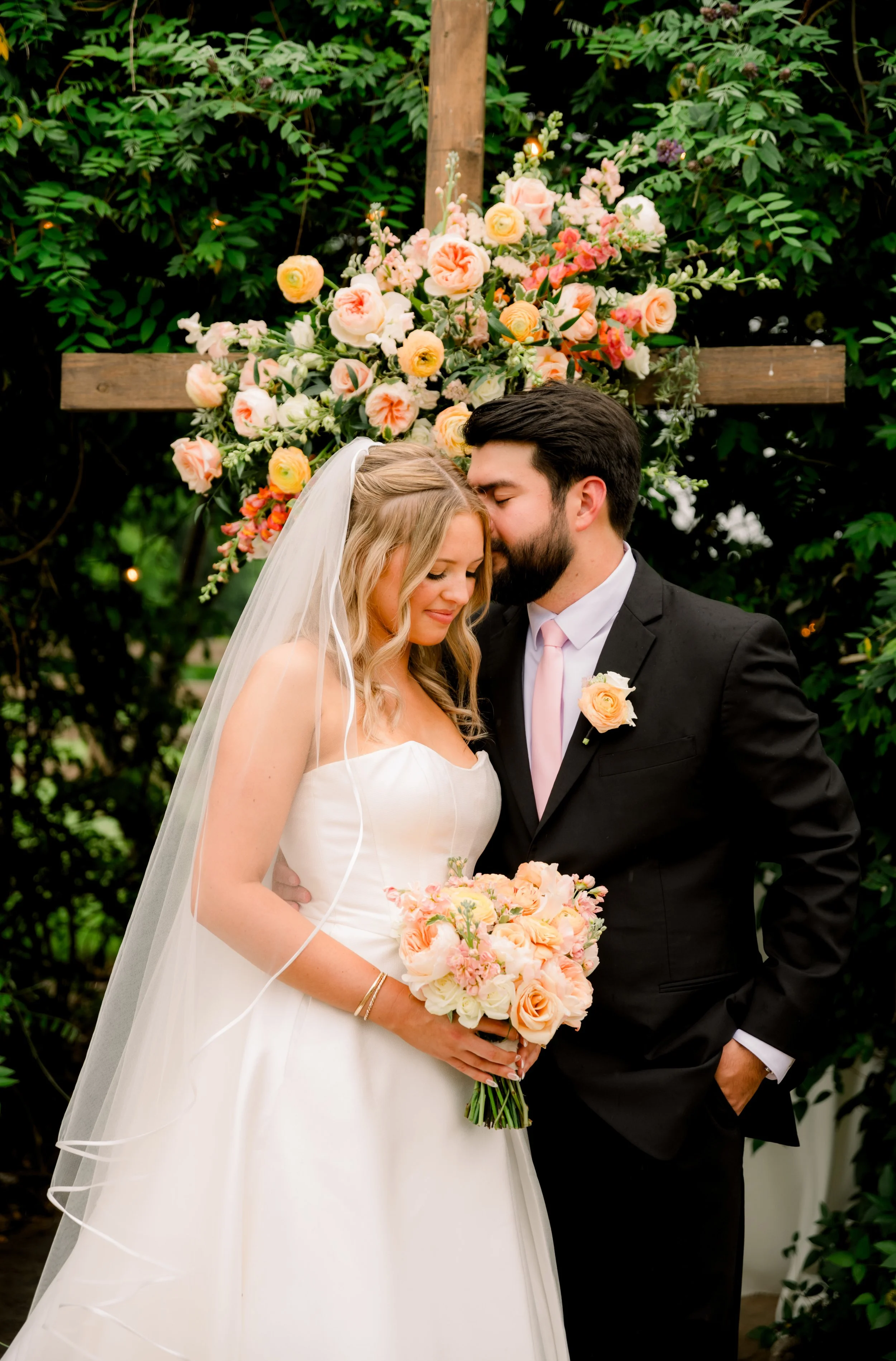 A bride and groom share a tender moment during their wedding ceremony, standing in front of a floral backdrop with a wooden cross, surrounded by greenery.