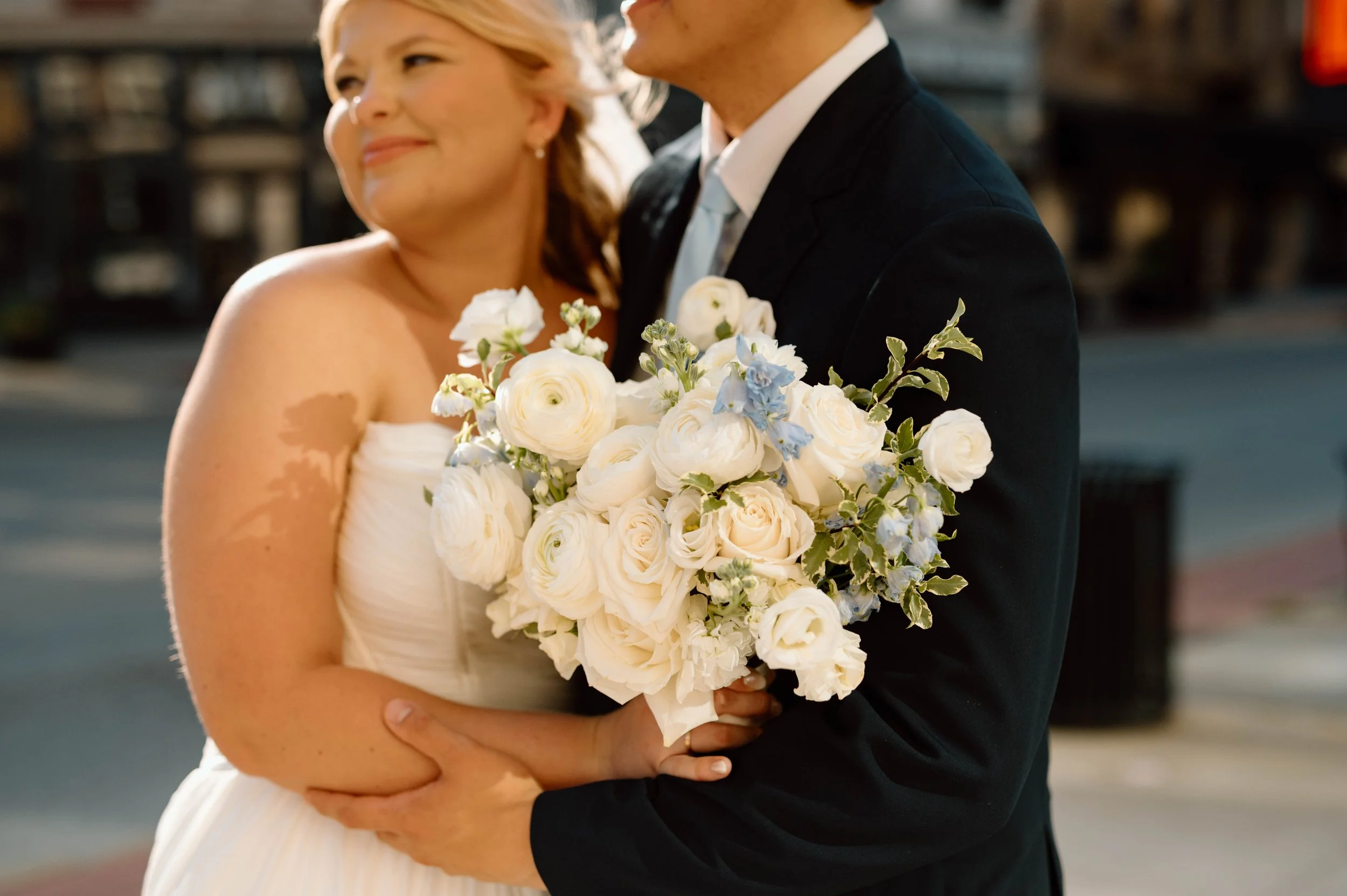 A bride in a white wedding dress holding a bouquet of white and light blue flowers, standing close to a groom in a black suit, outdoors during a wedding.
