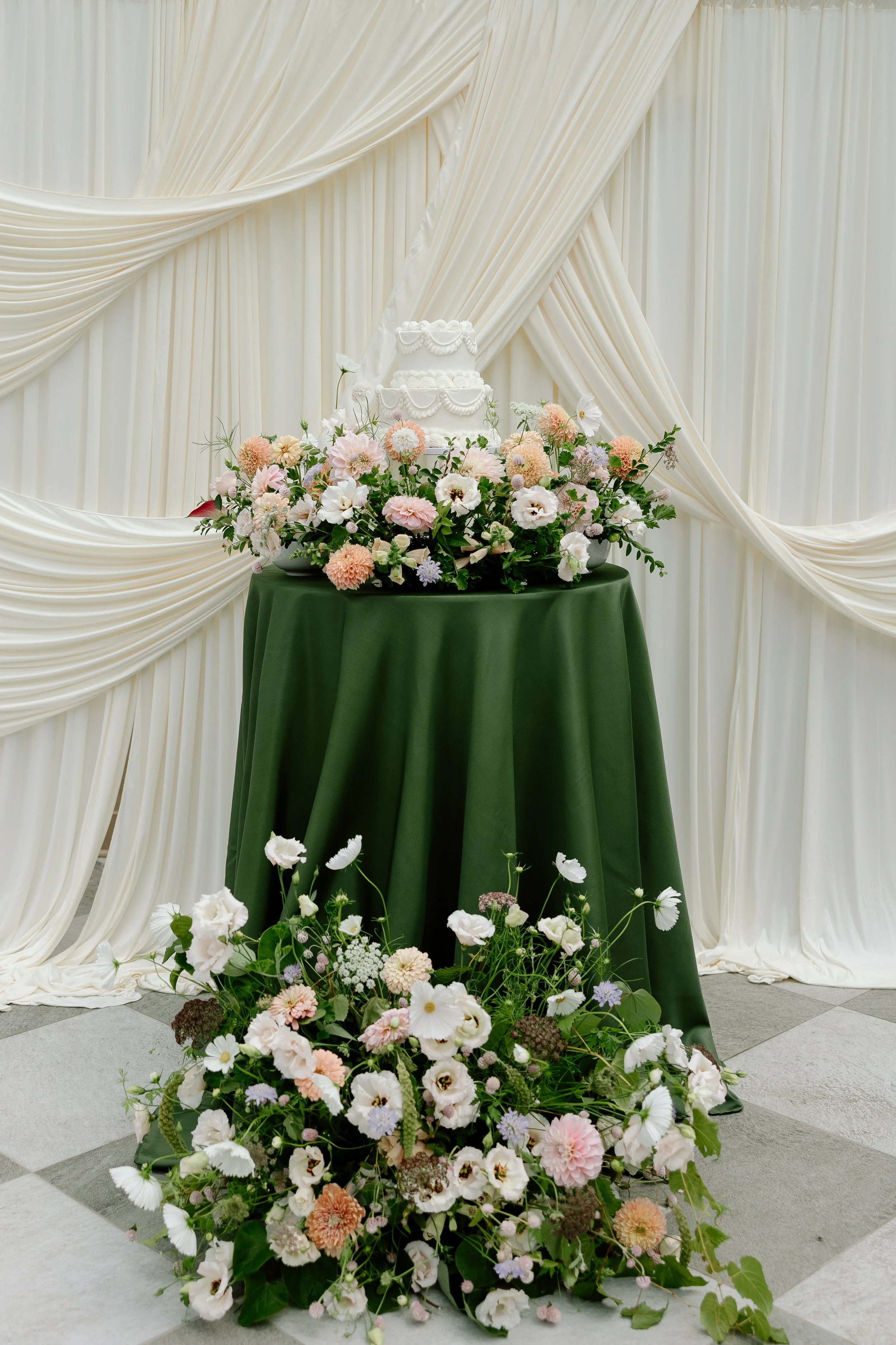 A white wedding cake with decorative piping, placed on a table covered with a green cloth, surrounded by pink and white flowers, with a white draped backdrop.