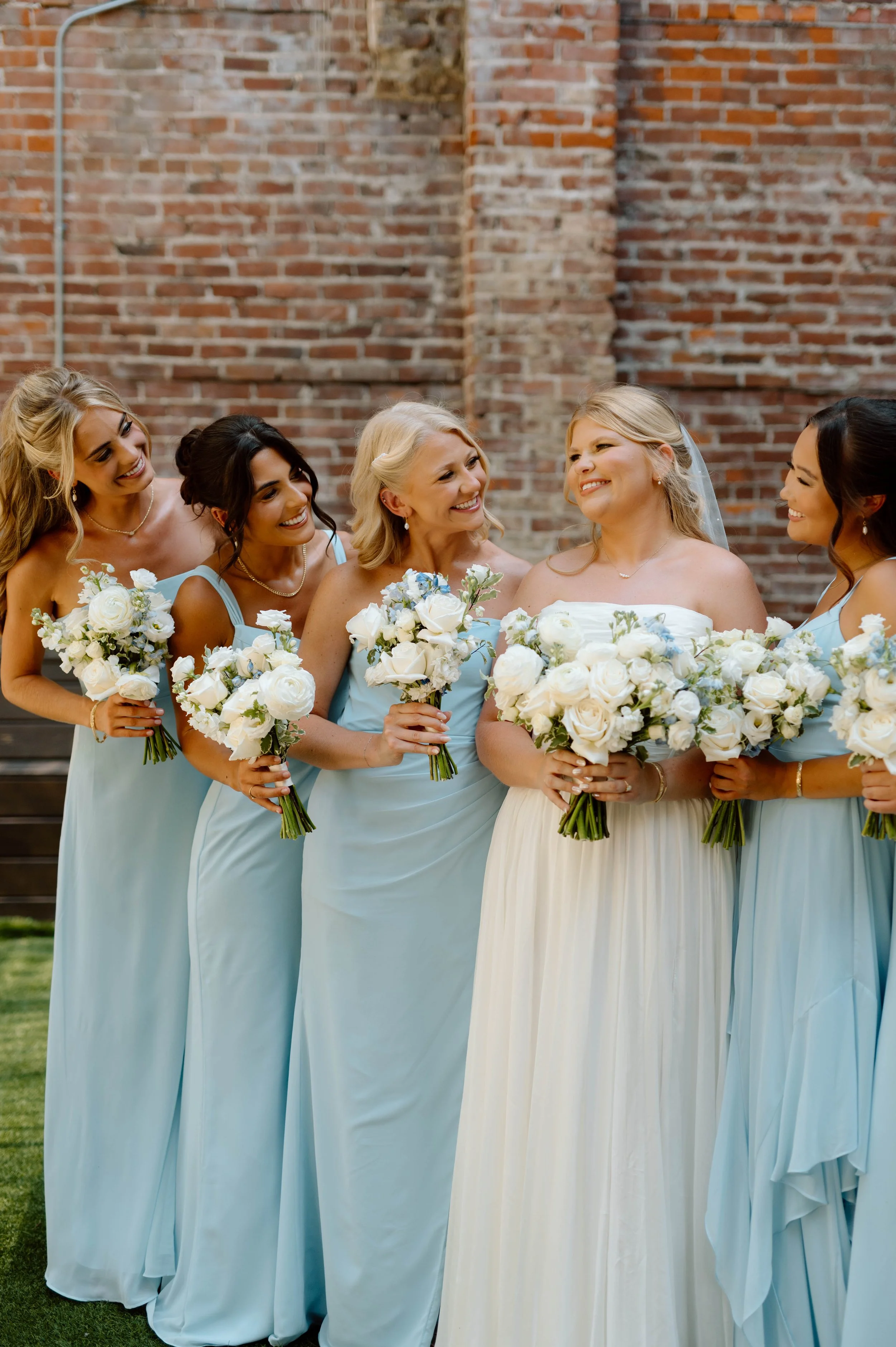 Bride and bridesmaids holding white bouquets, standing outside against a brick wall.