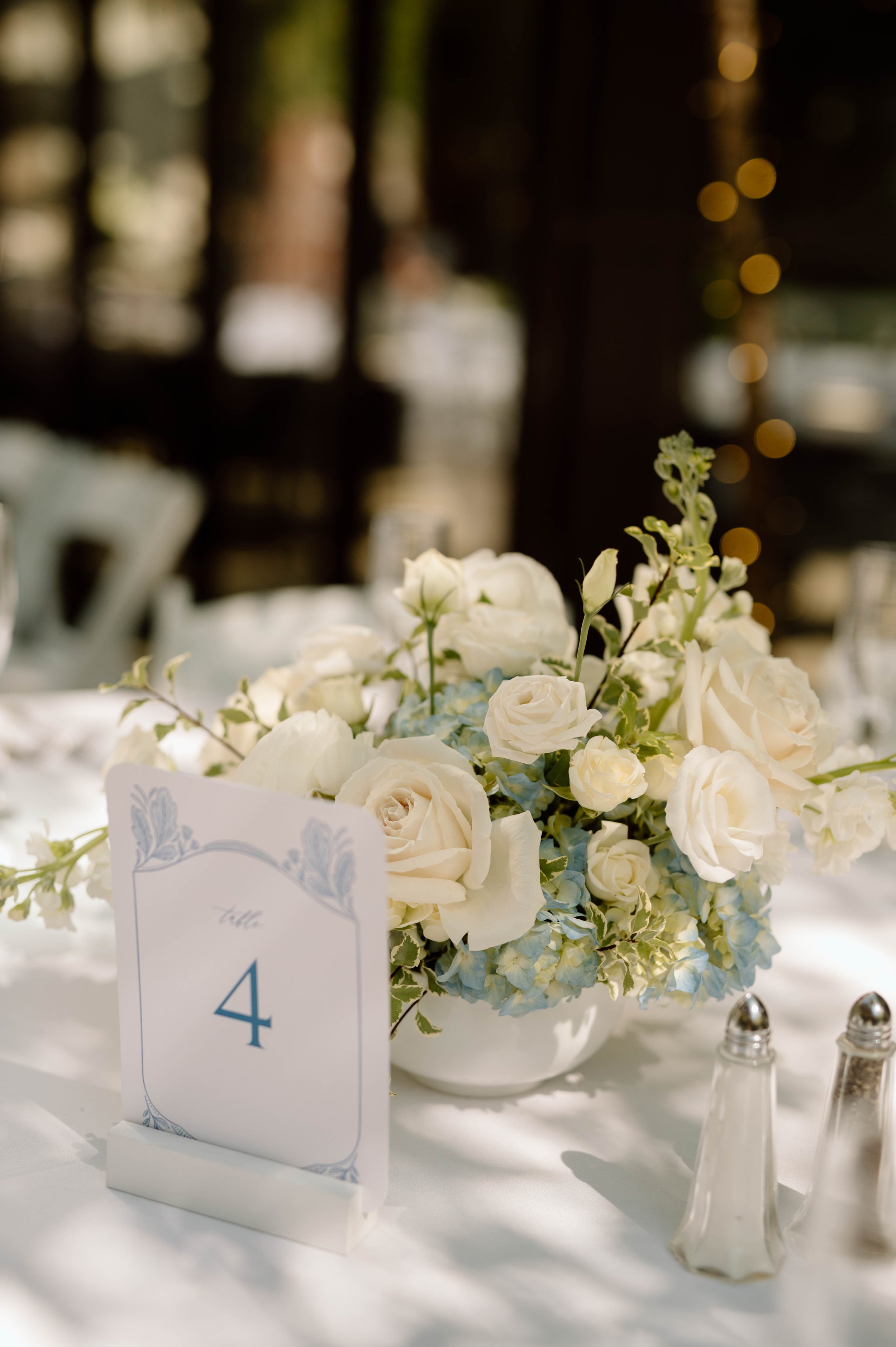 A table centerpiece with white roses and blue hydrangeas, a table number card with the number 4, and salt and pepper shakers.