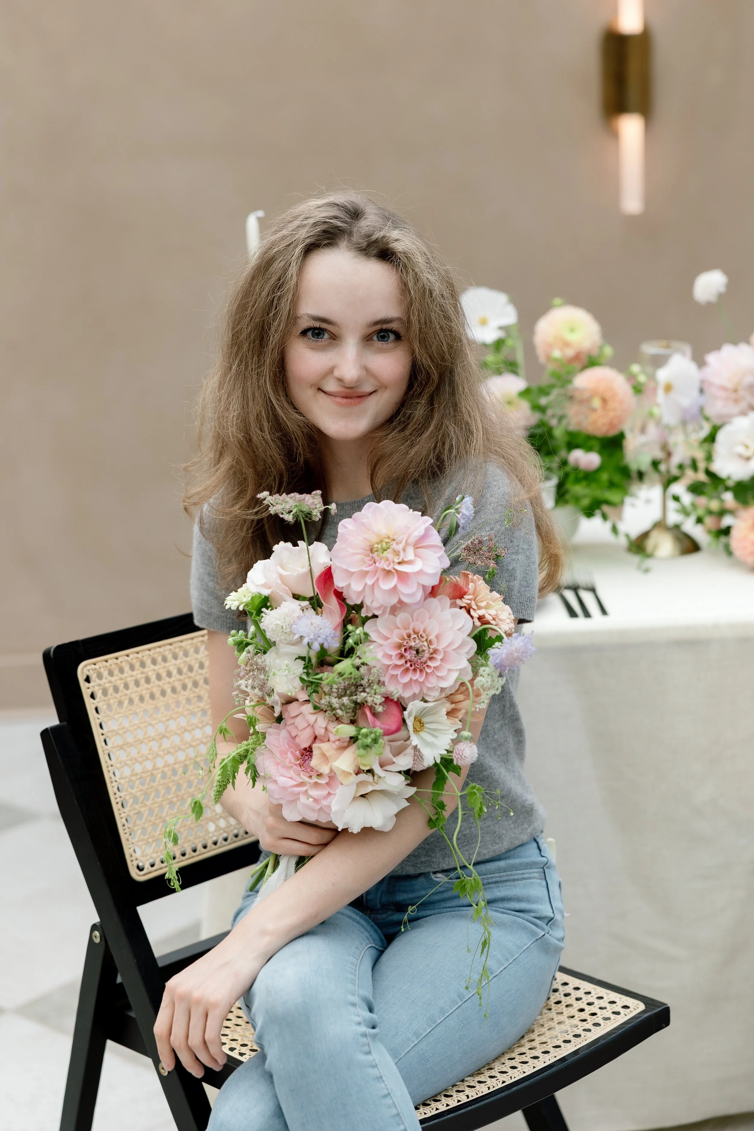A smiling young woman with long, wavy hair wearing a gray t-shirt and light blue jeans, holding a large bouquet of pink, white, and purple flowers, seated on a black chair with a wicker seat in an indoor setting decorated with more flowers in the background.