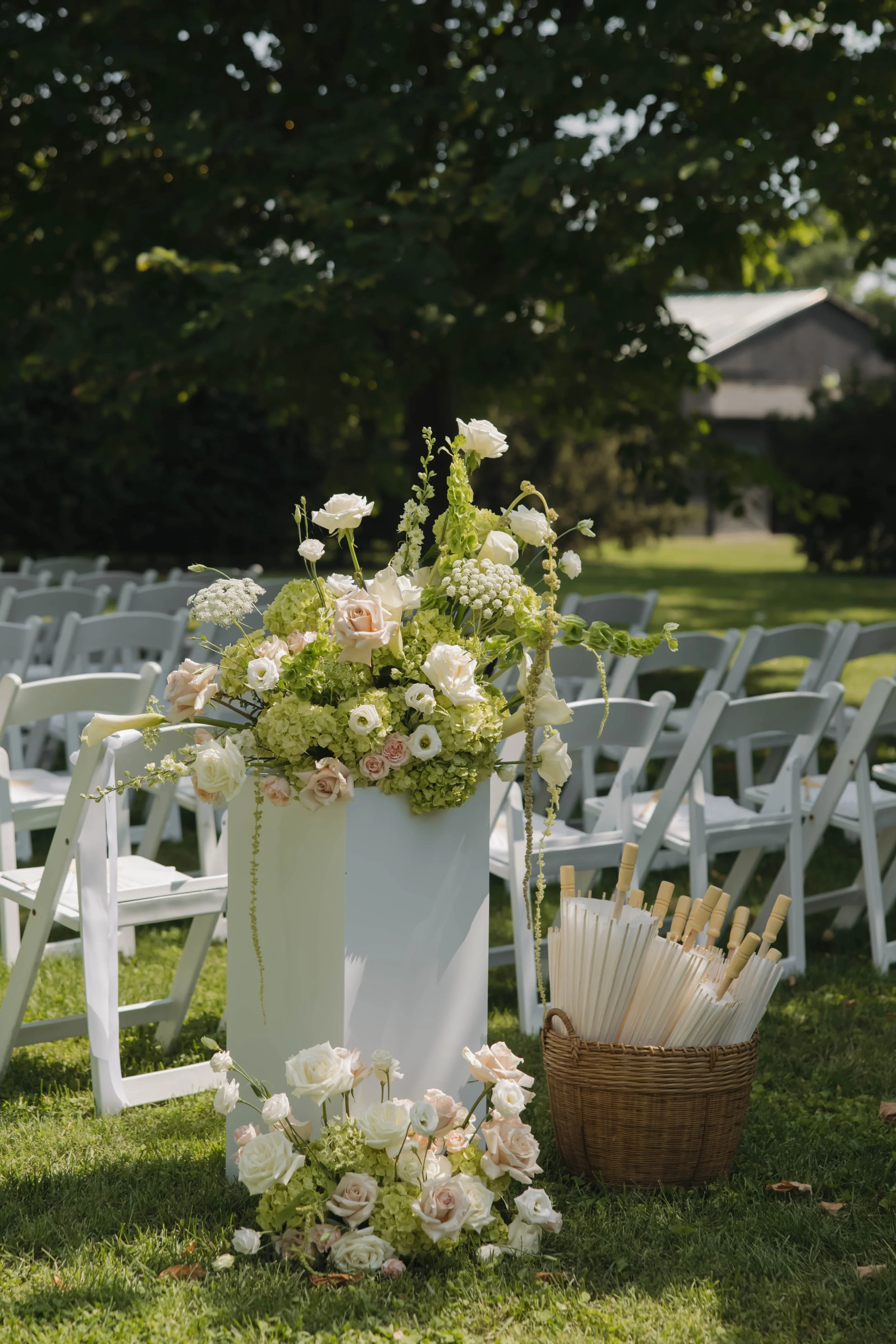 Flower arrangement and umbrellas at an outdoor wedding ceremony site with white chairs, a grassy area, and trees in the background.