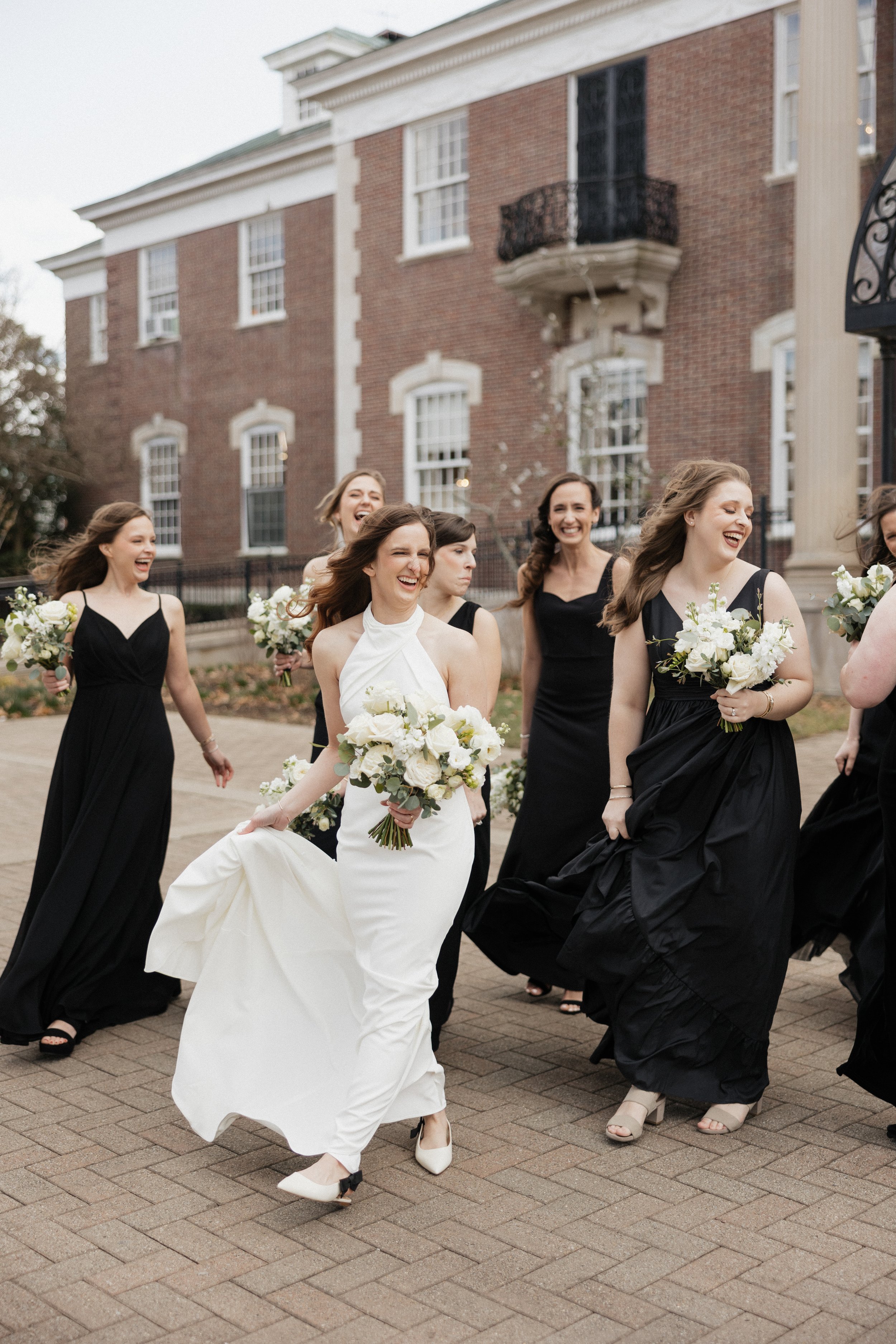 A group of women dressed in black and one in white wedding dress walking and smiling outside a brick building, holding bouquets of flowers.