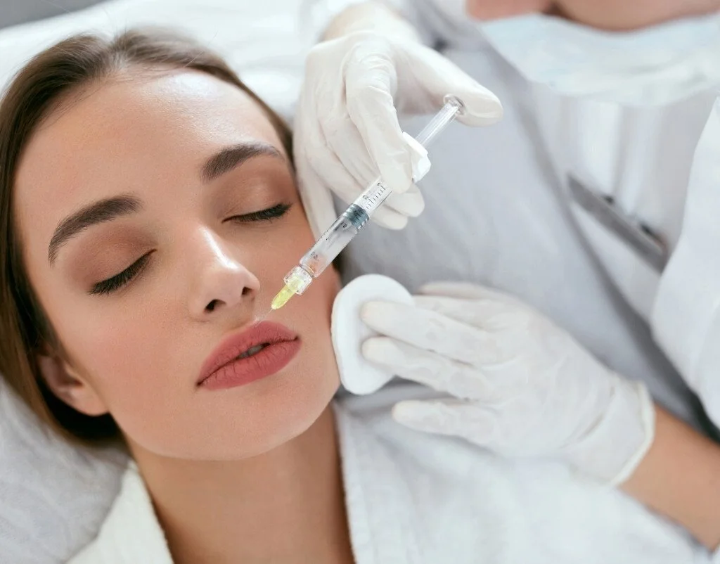 A woman receiving a cosmetic facial treatment from a medical professional holding a syringe and cotton pad.
