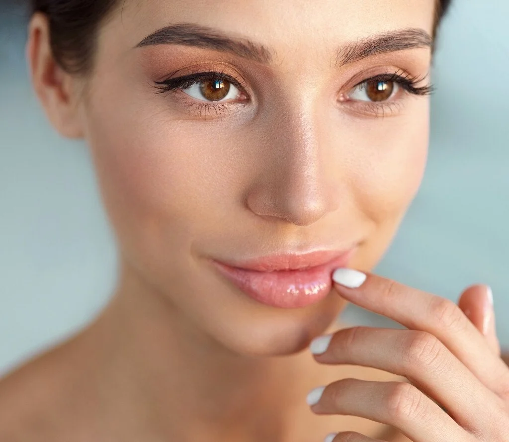 Close-up of a woman with light skin, brown eyes, and makeup, touching her lips with her fingers.
