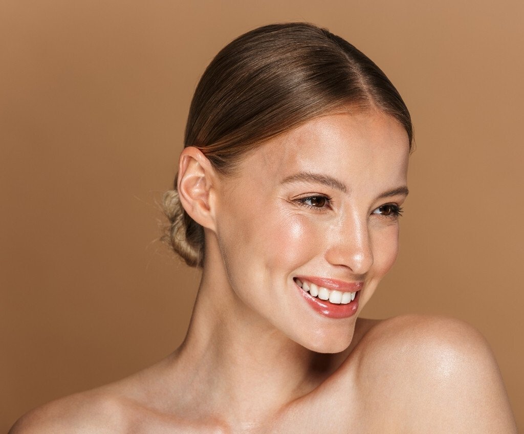 Close-up of a smiling young woman with clear skin and brown hair styled in a low bun, against a beige background.