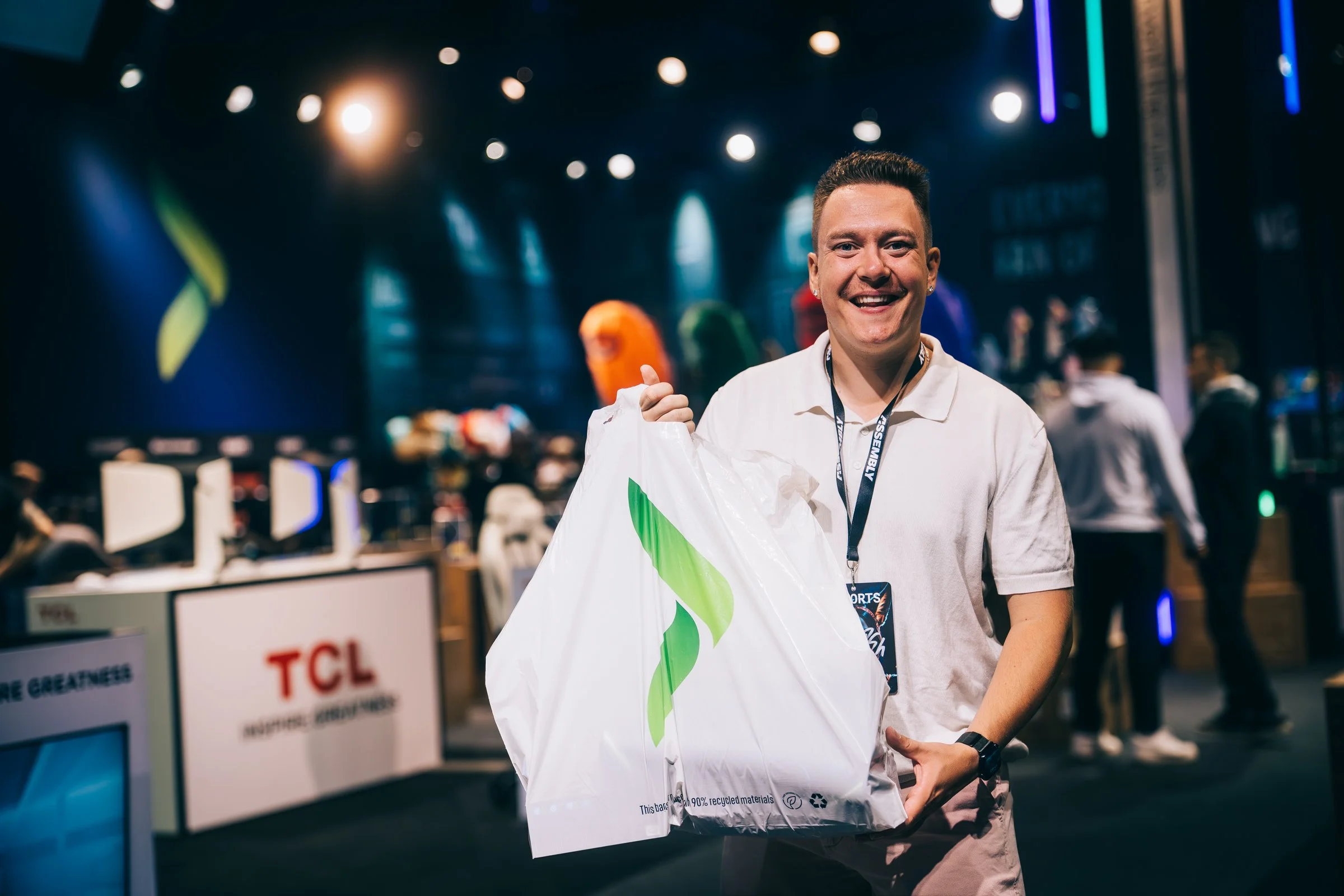 A smiling man holding a white shopping bag while standing in a busy indoor tech or gaming event with colorful lights and people in the background.