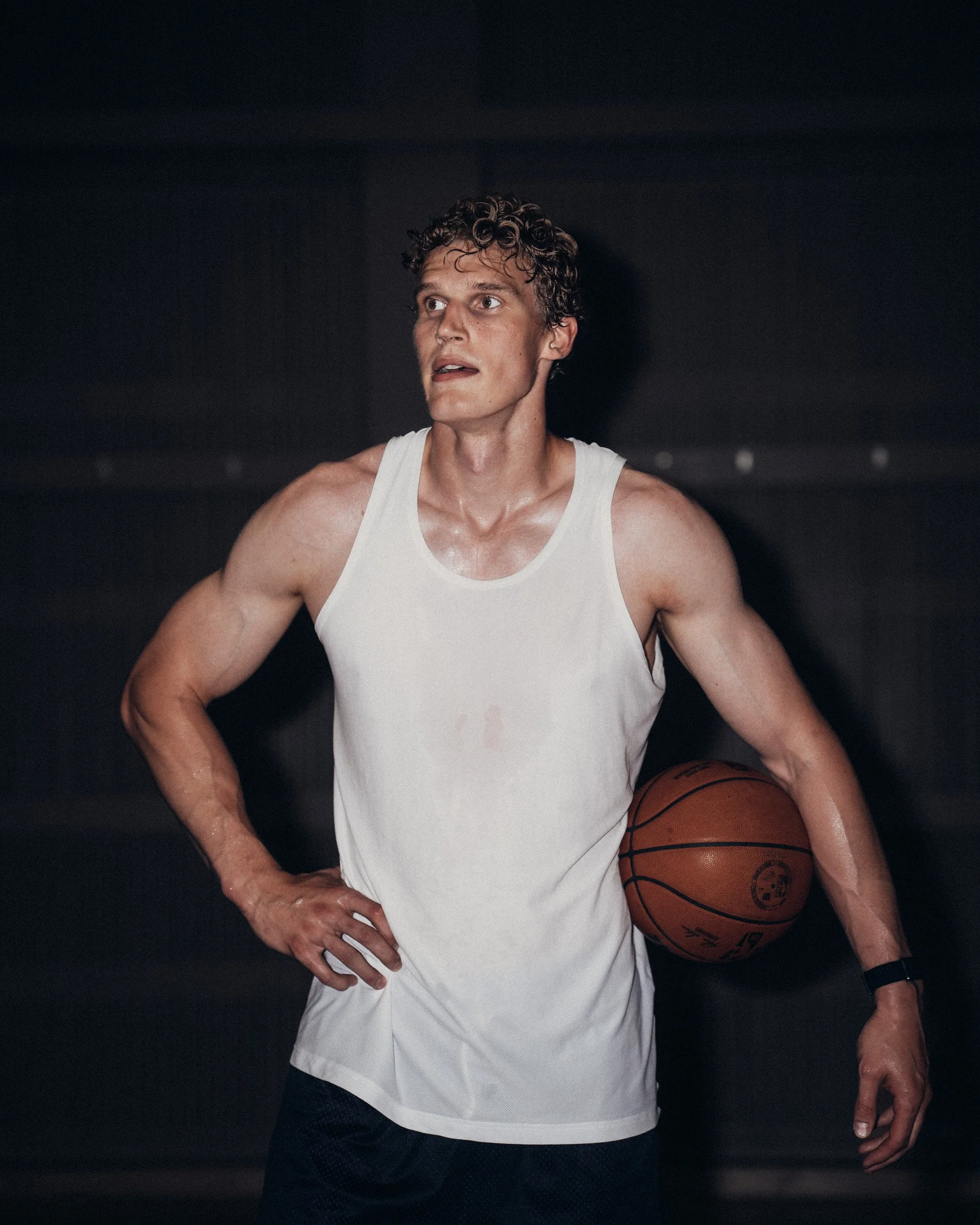 A young man in a white tank top holding a basketball, standing with one hand on his hip in a dark environment.