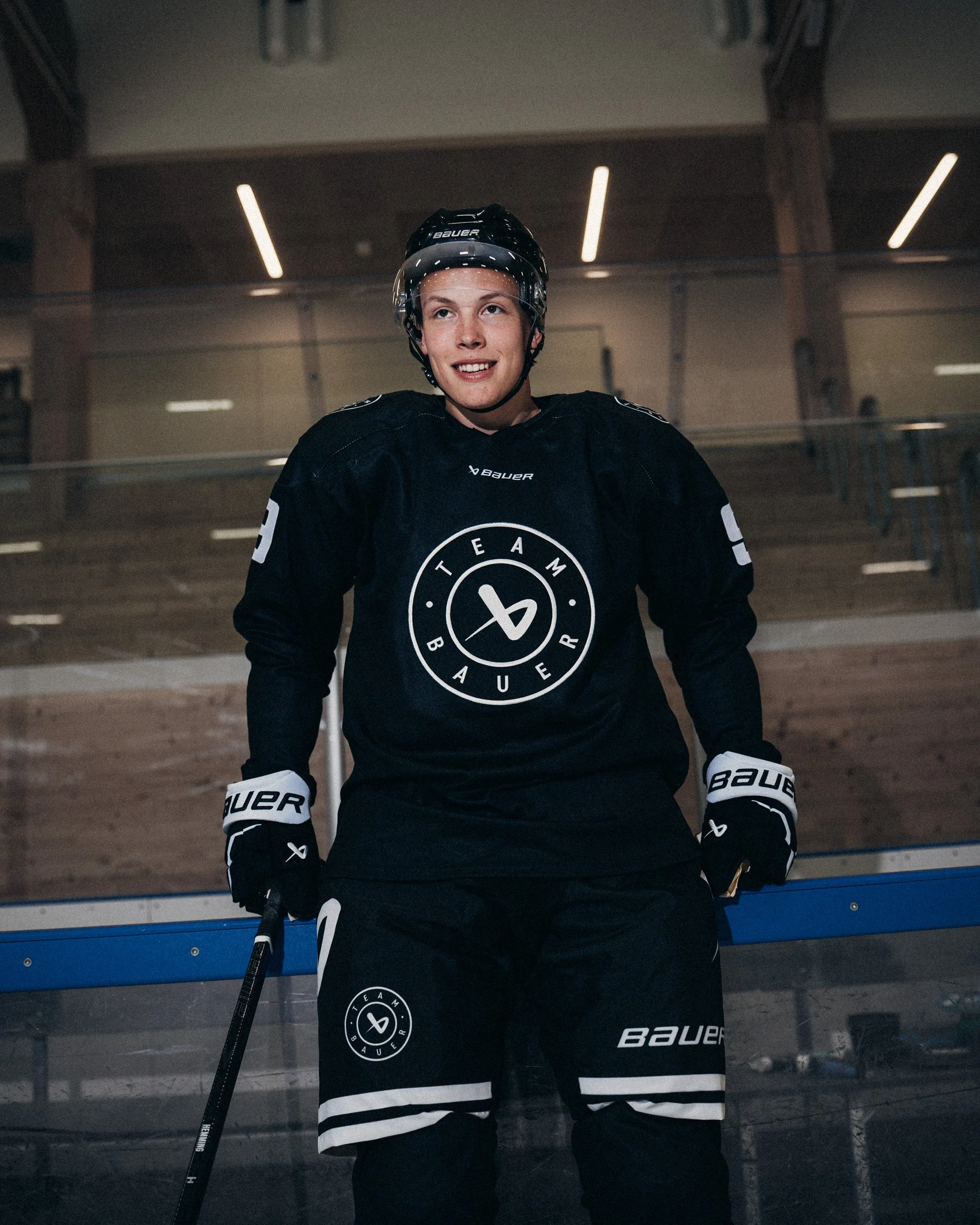 A young male hockey player wearing a helmet and black jersey with white accents stands on an ice rink, smiling. The jersey features the logo 'TEAM BAUER'.