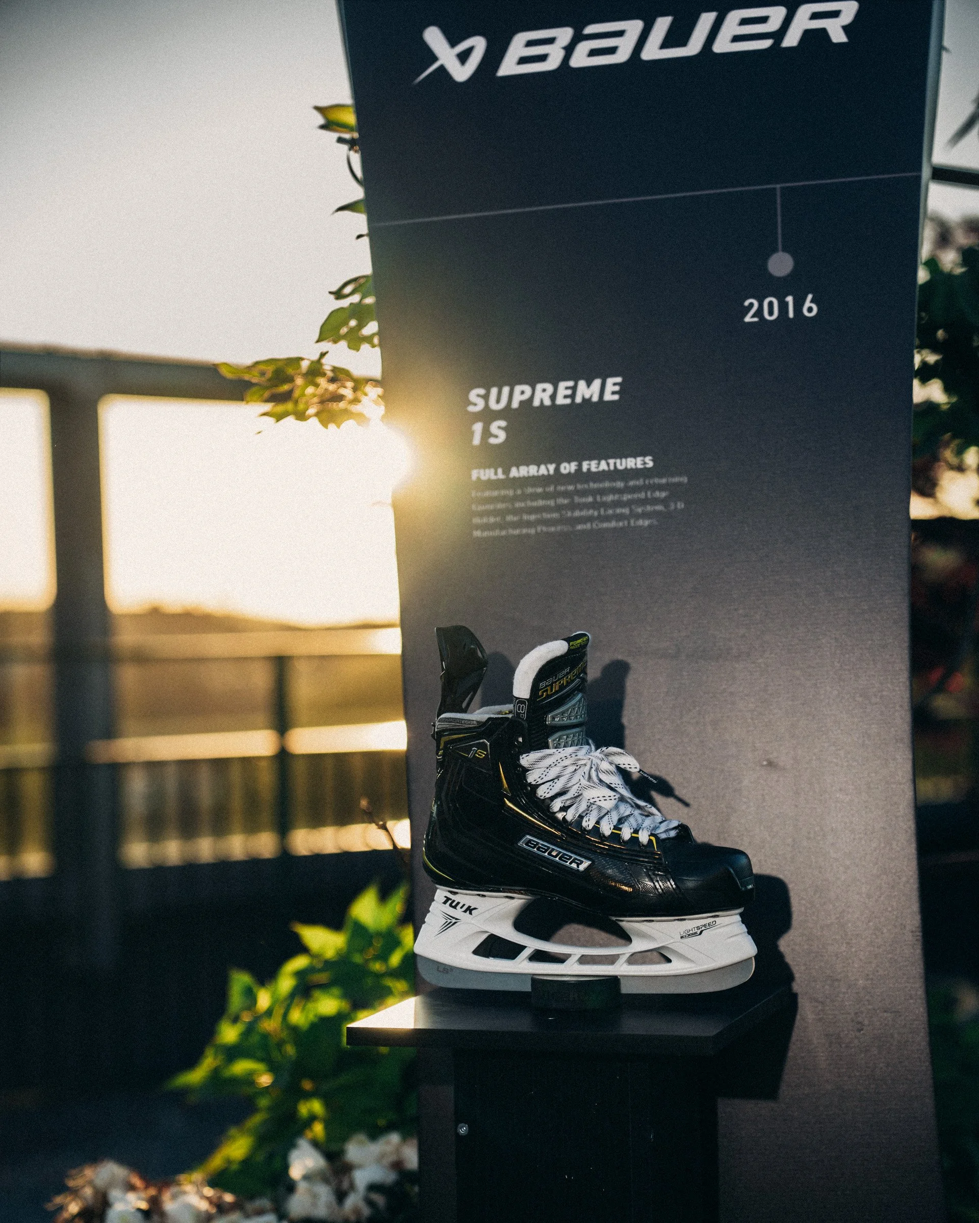 Display of a black hockey skate with white laces on a stand, next to a poster for Bauer Hockey featuring the text 'Supreme 1S' and a hockey stick, set against a background of trees and sunset.