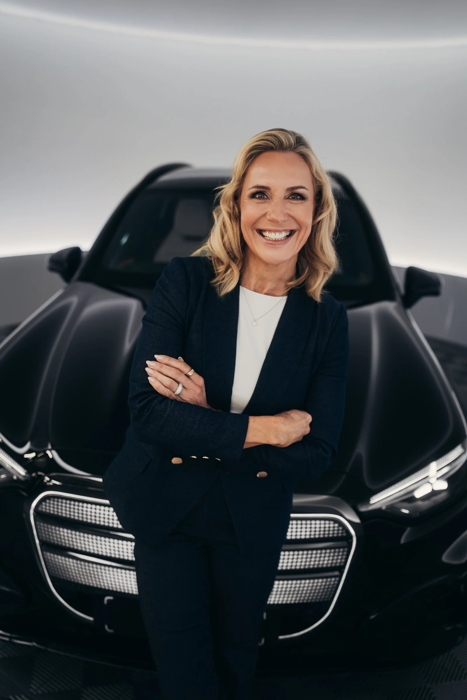 A woman with blonde hair smiling and crossing her arms, standing in front of a black luxury sports car.