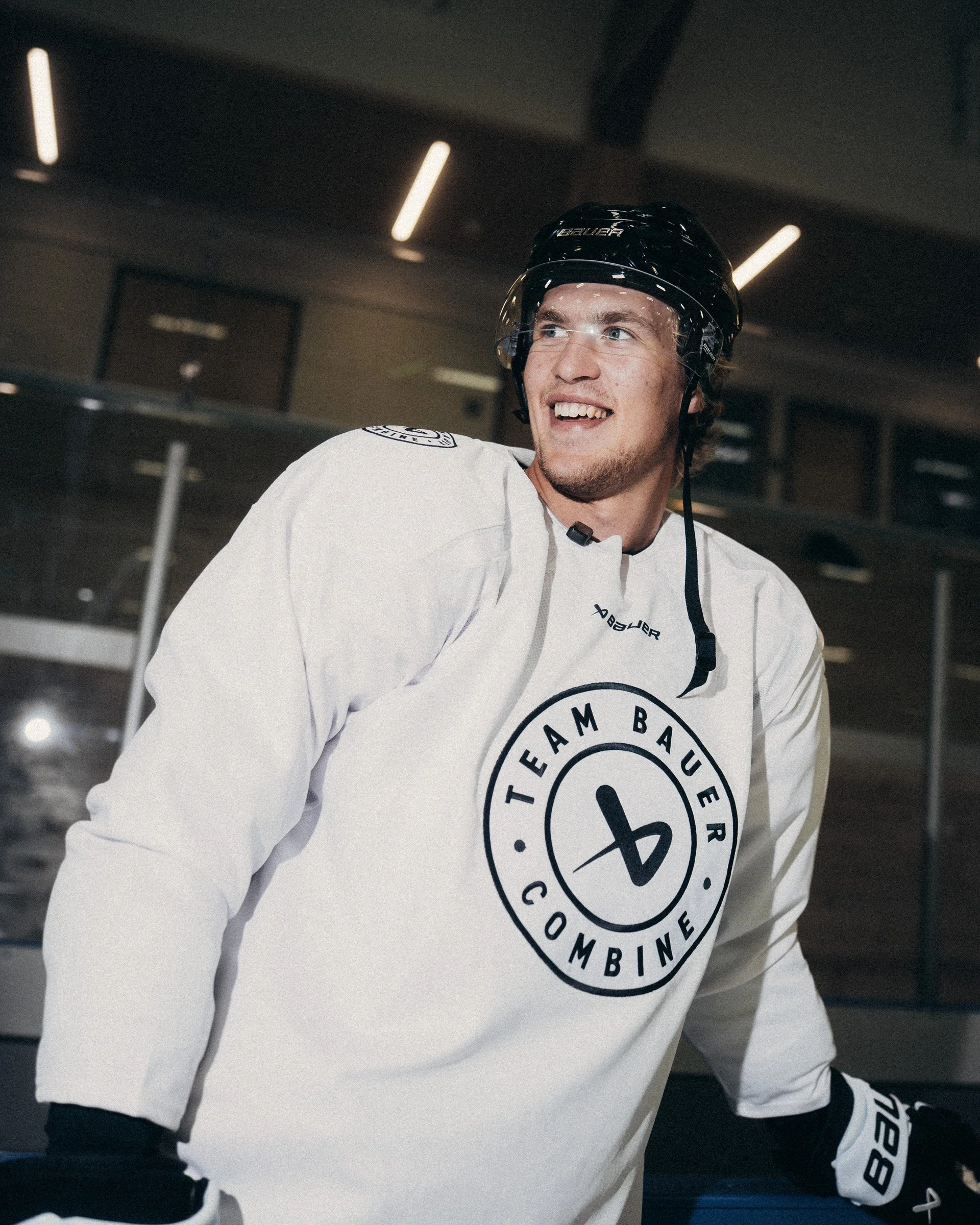 A young man in hockey gear, including a black helmet and white jersey with a team logo, smiling in an indoor ice rink.