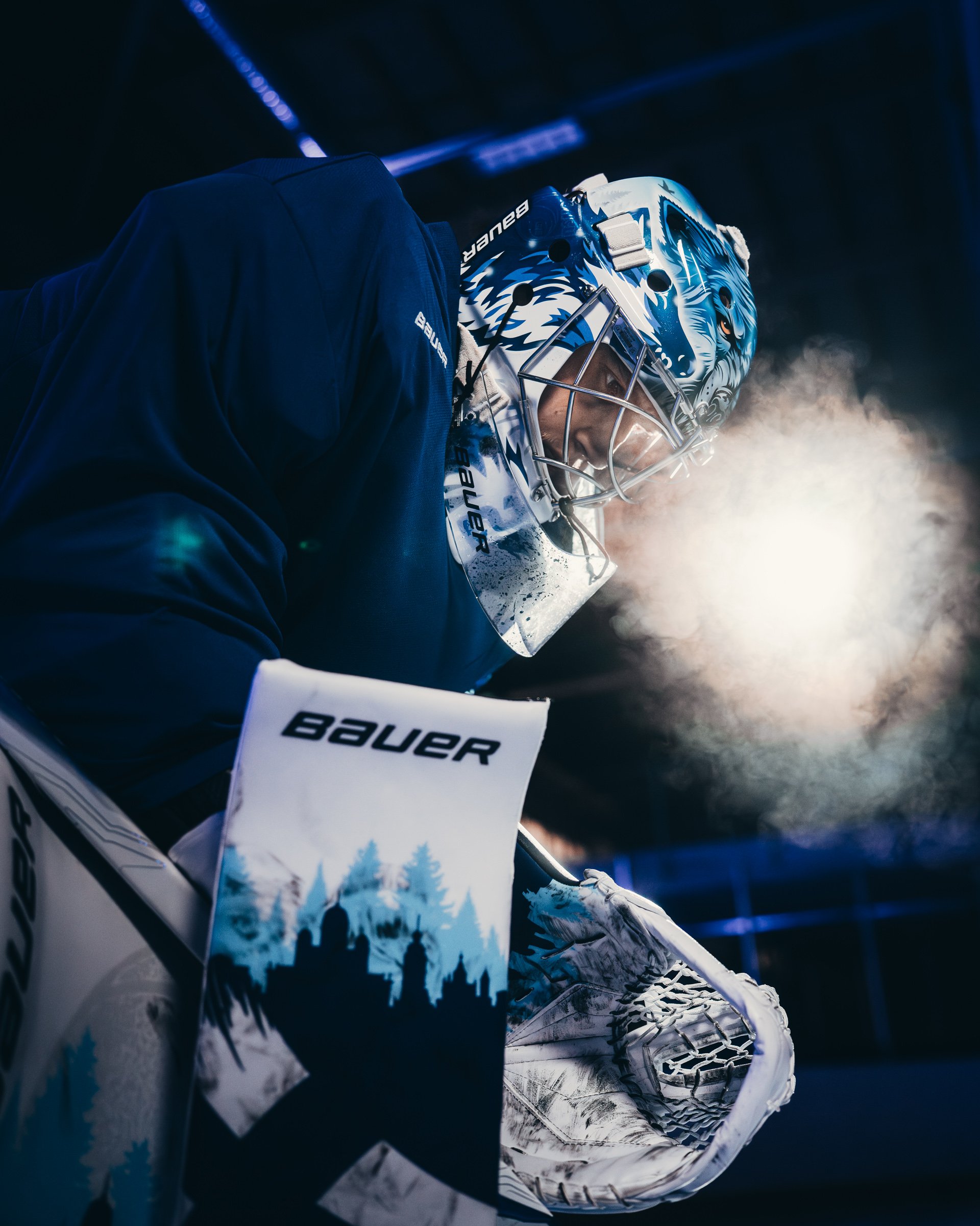 Close-up of an ice hockey goalie in a blue jersey and a helmet with a blue and white wolf design, standing in a dimly lit arena with visible smoke or mist.