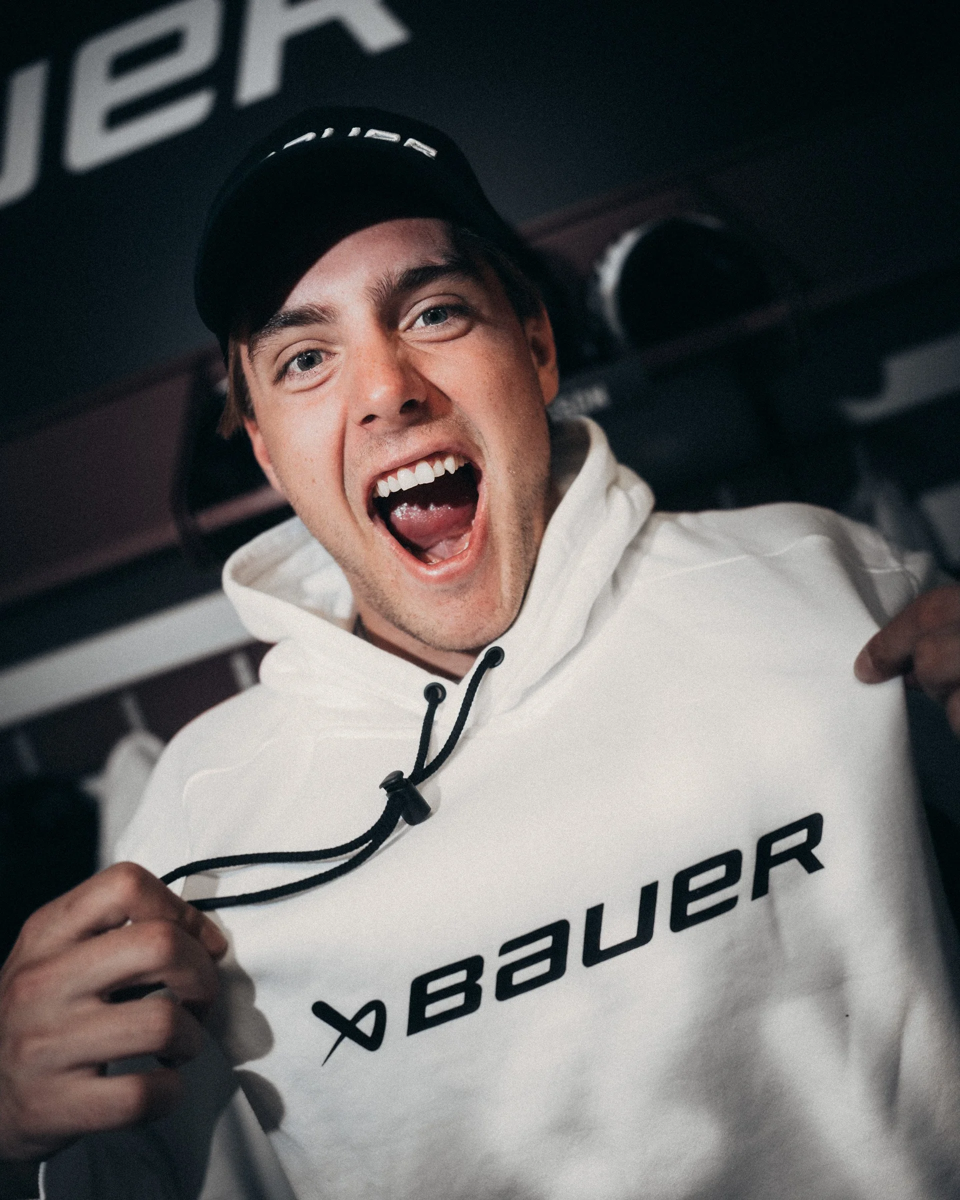 A young man is smiling widely, showing his teeth, and wearing a black cap and a white hoodie. He appears excited or happy, and is holding some object or part of a sign in front of him. The background has large printed text, possibly part of a billboa