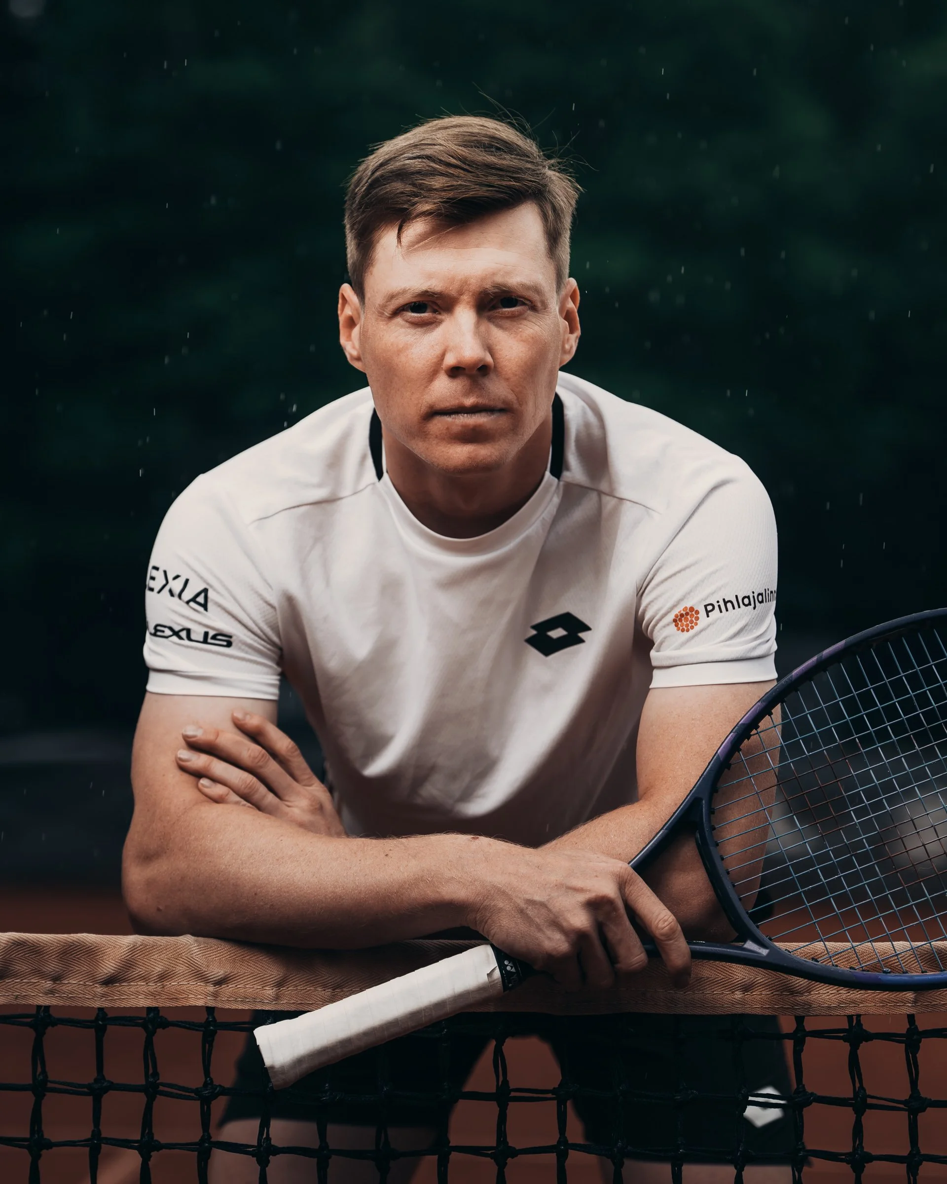A male tennis player sitting on a tennis court holding a tennis racket, wearing a white sports shirt with logos, and resting his arm on a net.