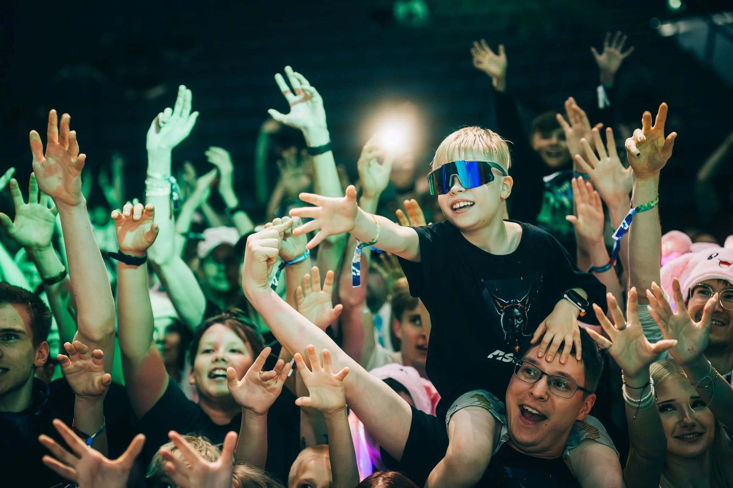 A young boy wearing sunglasses is lifted above a crowd at a concert or event, with many raised hands and smiling faces around him.
