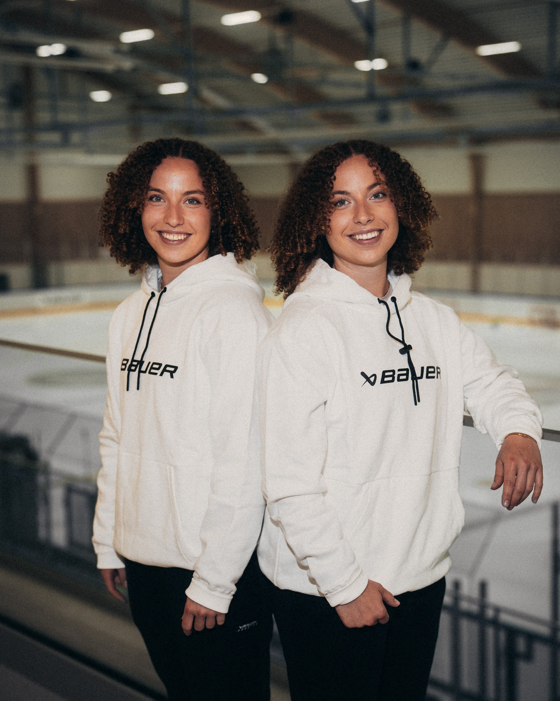 Two women with curly hair smiling and standing back-to-back at an indoor ice rink, wearing white hoodies and black pants.